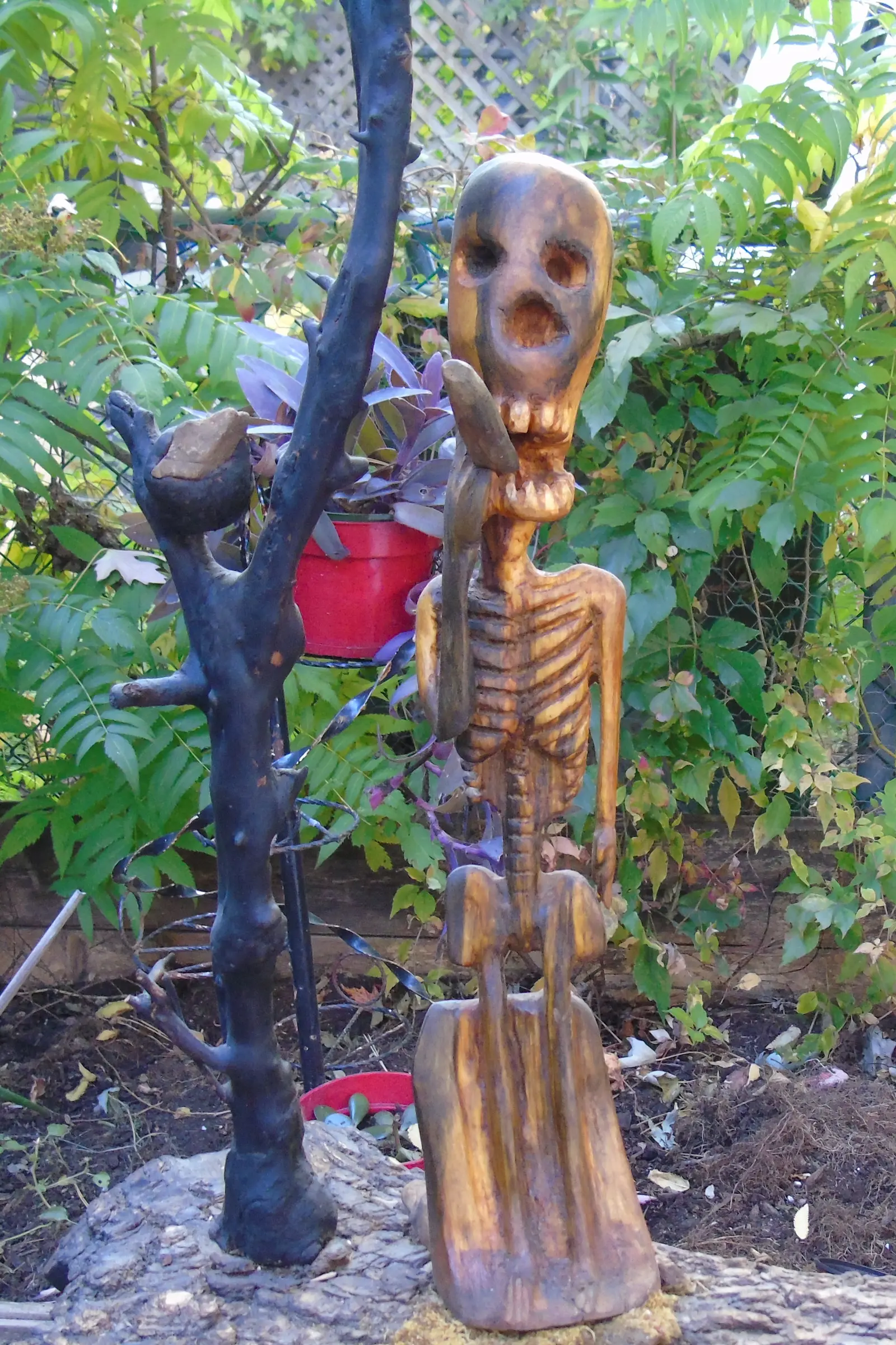 Mort mais vivant 1 : vue de face, la figure squelettique en bois clair debout sur son socle, une pierre tenue contre la mâchoire, la branche calcinée dressée à gauche, les deux sur une plaque d'écorce garnie de mousse, devant un feuillage dense
