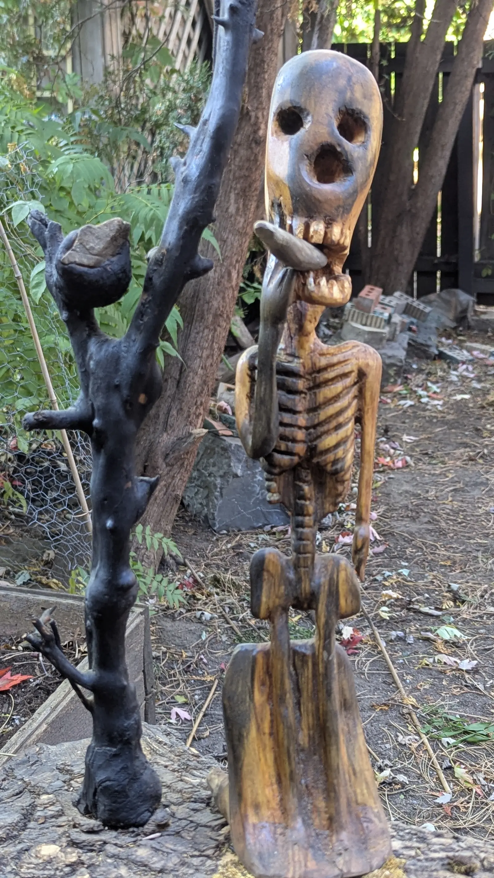 Mort mais vivant 1 : les deux verticales de plus près, le crâne aux orbites rondes et à la bouche ouverte, les côtes en relief, le socle en coin rayé de brun, la branche noire et sa pierre, devant une clôture et des briques