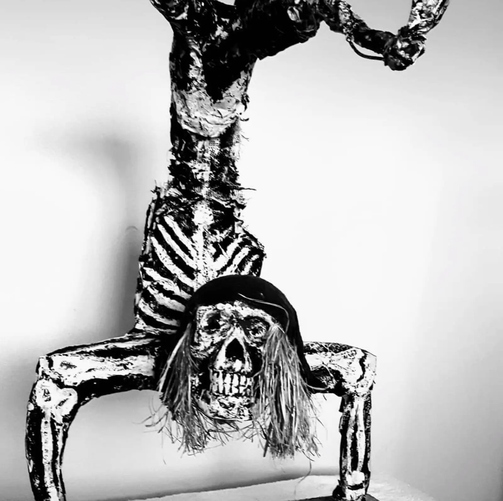Skeleton balancing on its hands, body caked in a crusted coating with gauze along the spine, a dark cap and long pale fibres on the skull, standing against a white wall in high-contrast black and white