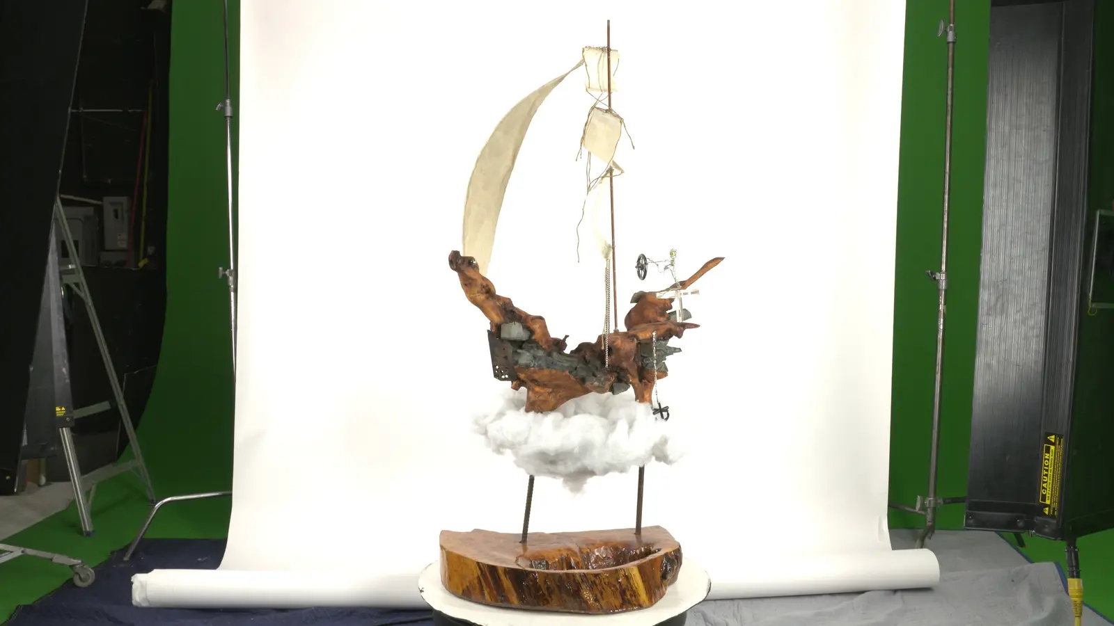 Coque en bois trouvé sculpté + voile en tissu + mât avec voilures + petits squelettes en bois sur le pont, suspendu sur nuages d'ouate, socle en rondin. — vue 1