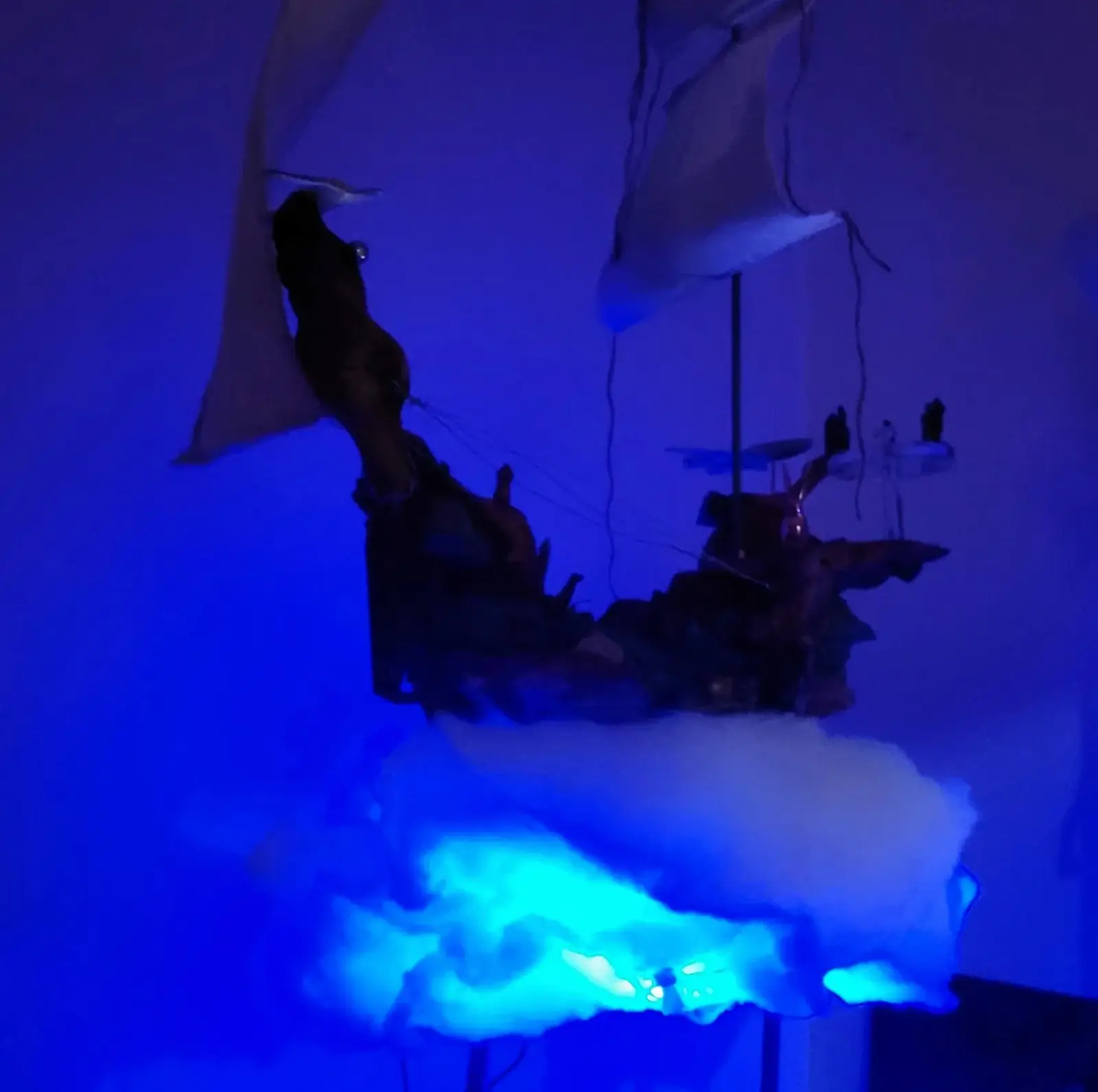 Vue du voilier en bois trouvé dans une pièce sombre, baigné de lumière bleue, les voiles de tissu pâle dressées au-dessus de la coque et le nuage d'ouate illuminé par en dessous, socle hors cadre
