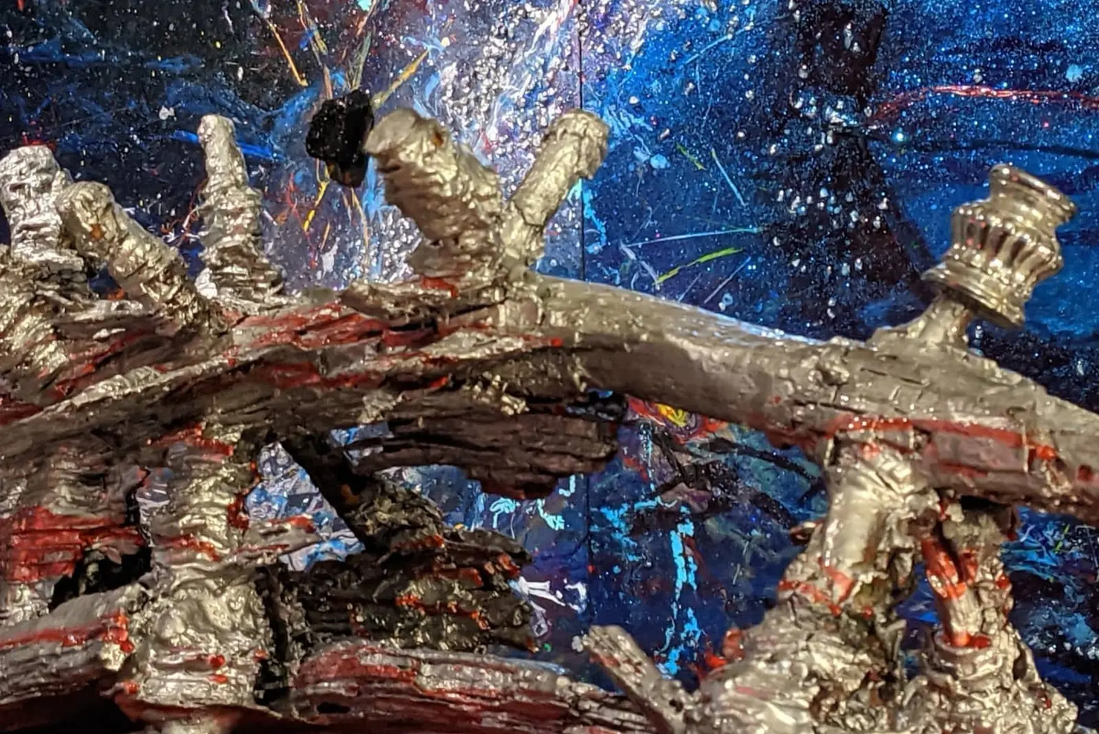 Gros plan sur les crêtes argentées du bois, des traces rouges au fond des sillons, un petit élément chromé tourné à droite, devant une peinture bleu nuit éclaboussée de blanc