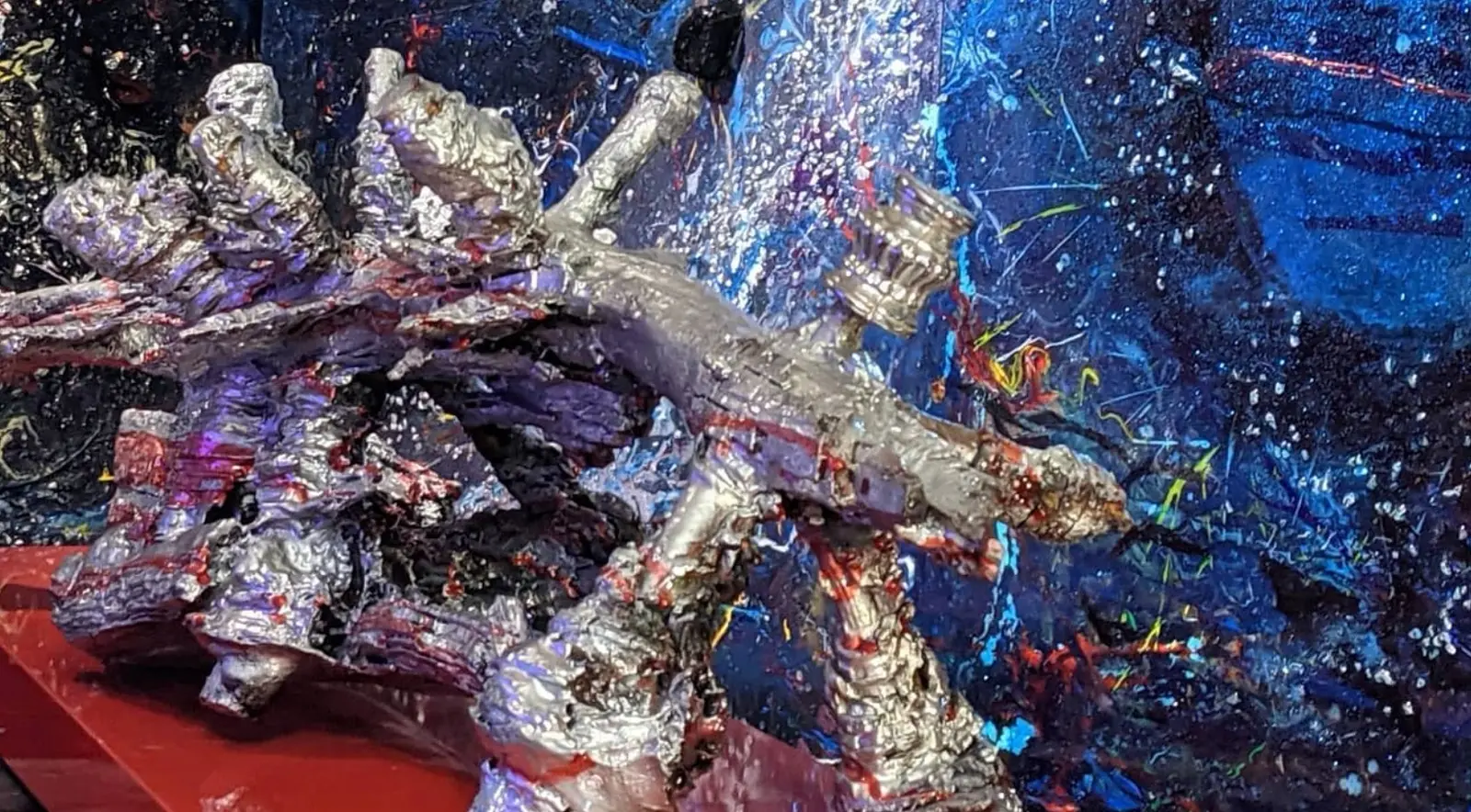 Vue de trois quarts en plongée, la masse argentée posée sur un plateau rouge laqué, ses moignons dressés vers le haut, devant une peinture bleue griffée de blanc et de couleurs vives