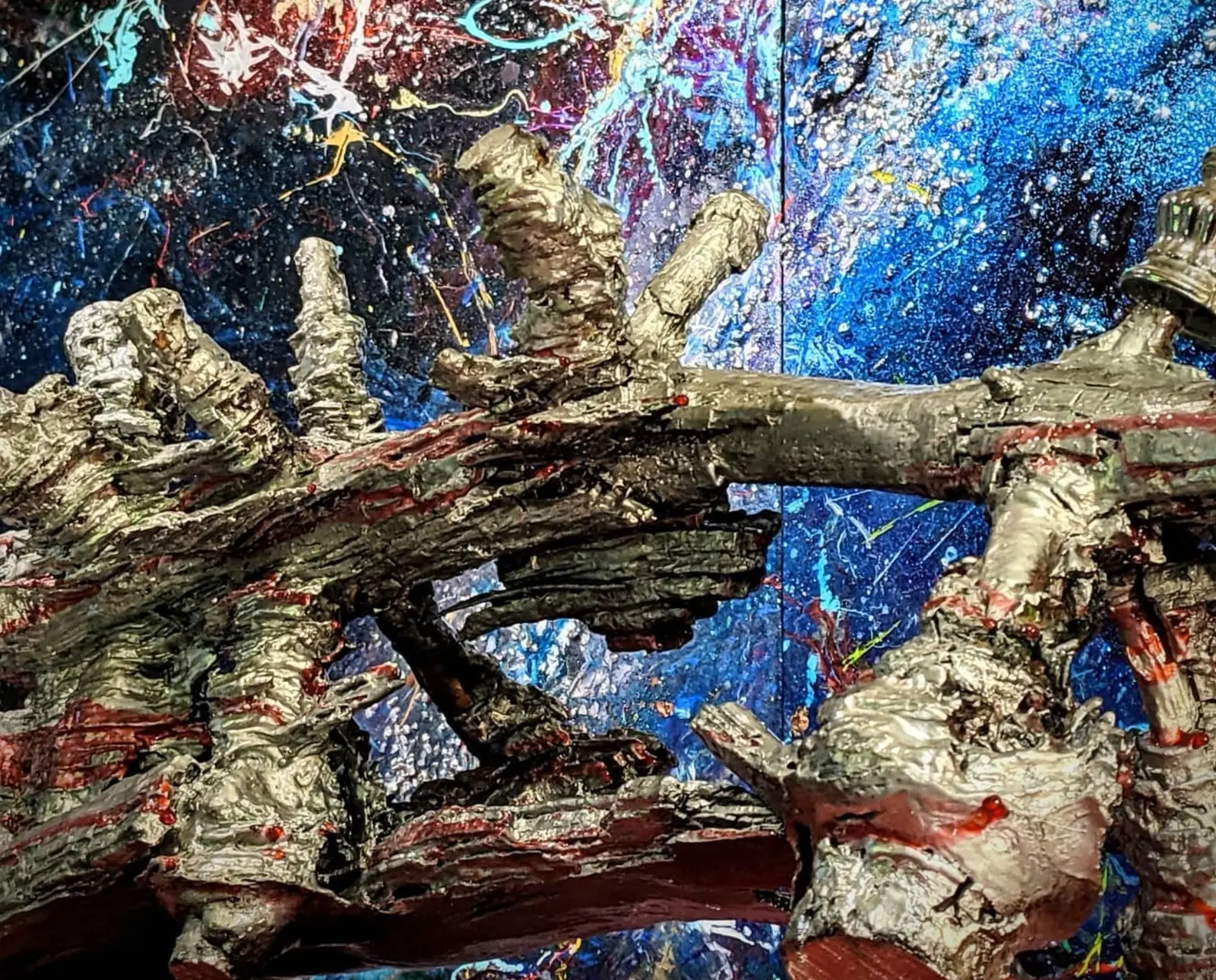 Cadrage rapproché sur la partie haute, la longue branche argentée enjambe une cavité sombre, l'écorce rougeâtre apparaît sous le métal, et derrière, la jointure entre deux toiles bleues constellées
