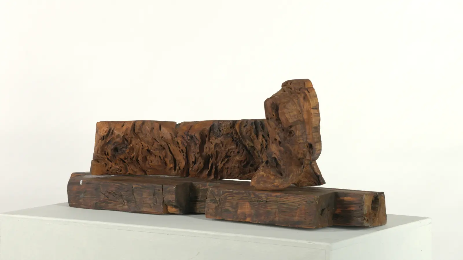 Vue de trois quarts de la figure couchée, masse de loupe sombre posée sur les poutres récupérées, tête à droite, fond blanc