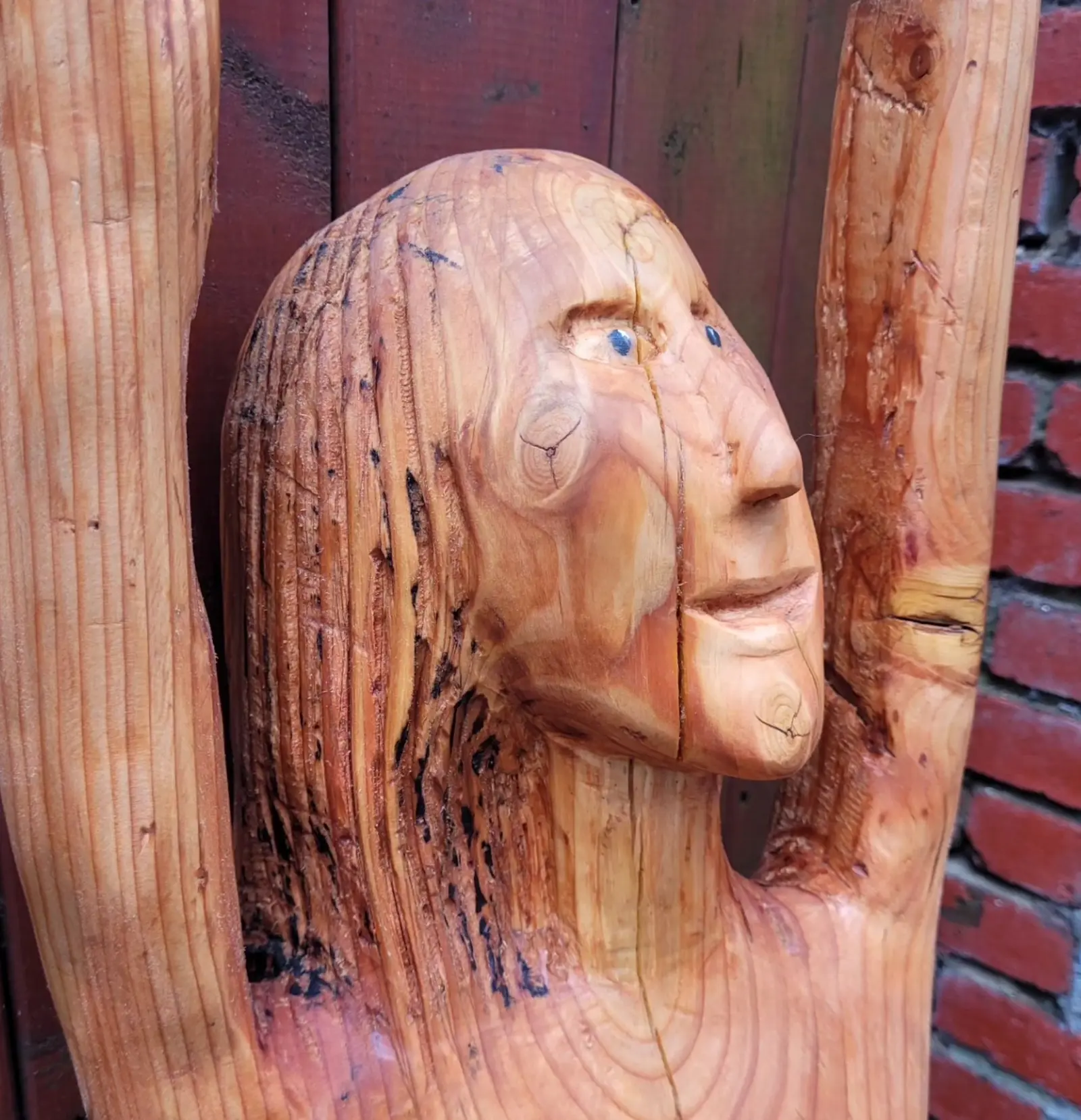 Close-up of the head turned three-quarters, hair carved into long strands streaked with black, two small dark blue beads set into the eyes, and the raised arms framing the face against the fence and brick wall