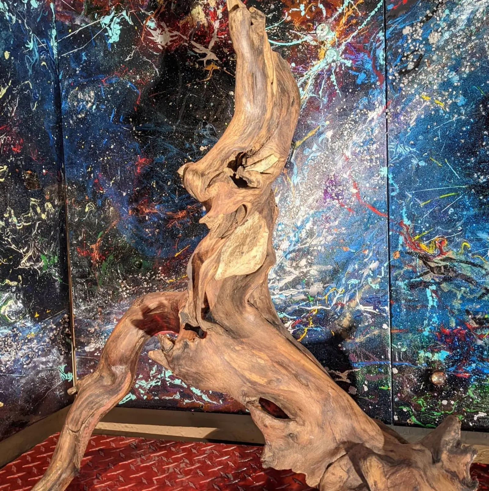 Vue rapprochée en contre-plongée, le bois gris beige et mat, deux pierres claires logées dans les cavités, l'œuvre posée sur un sol de tôle rouge dans un angle occupé par deux grands tableaux éclaboussés de couleurs