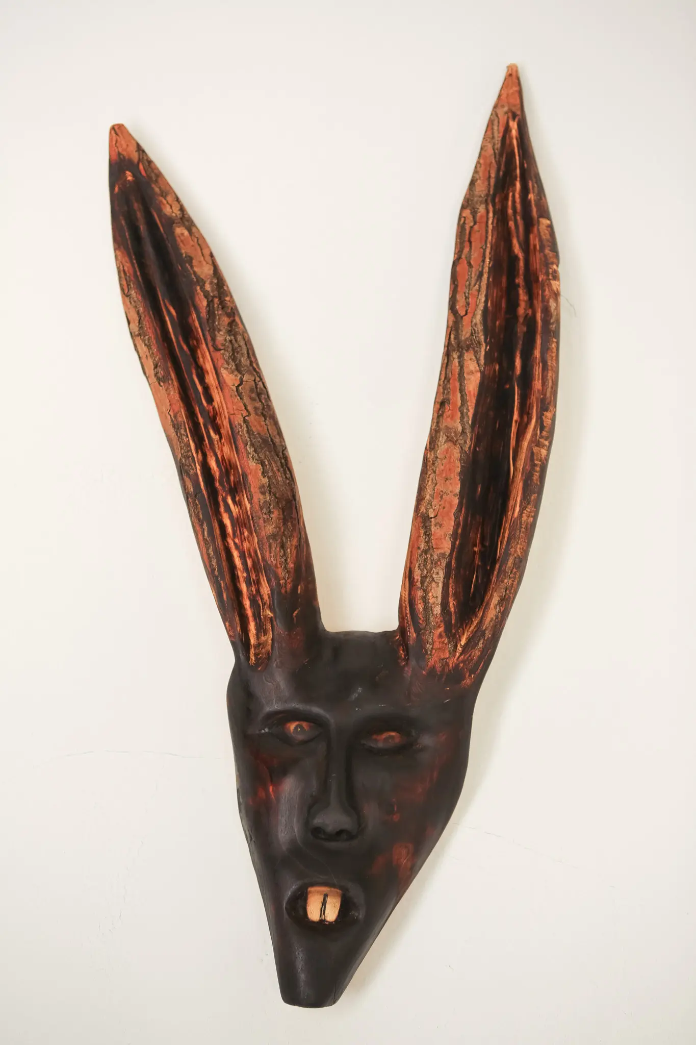Masque mural en bois trouvé, face léporine assombrie et deux longues oreilles dressées à l'écorce orangée, vue de face.