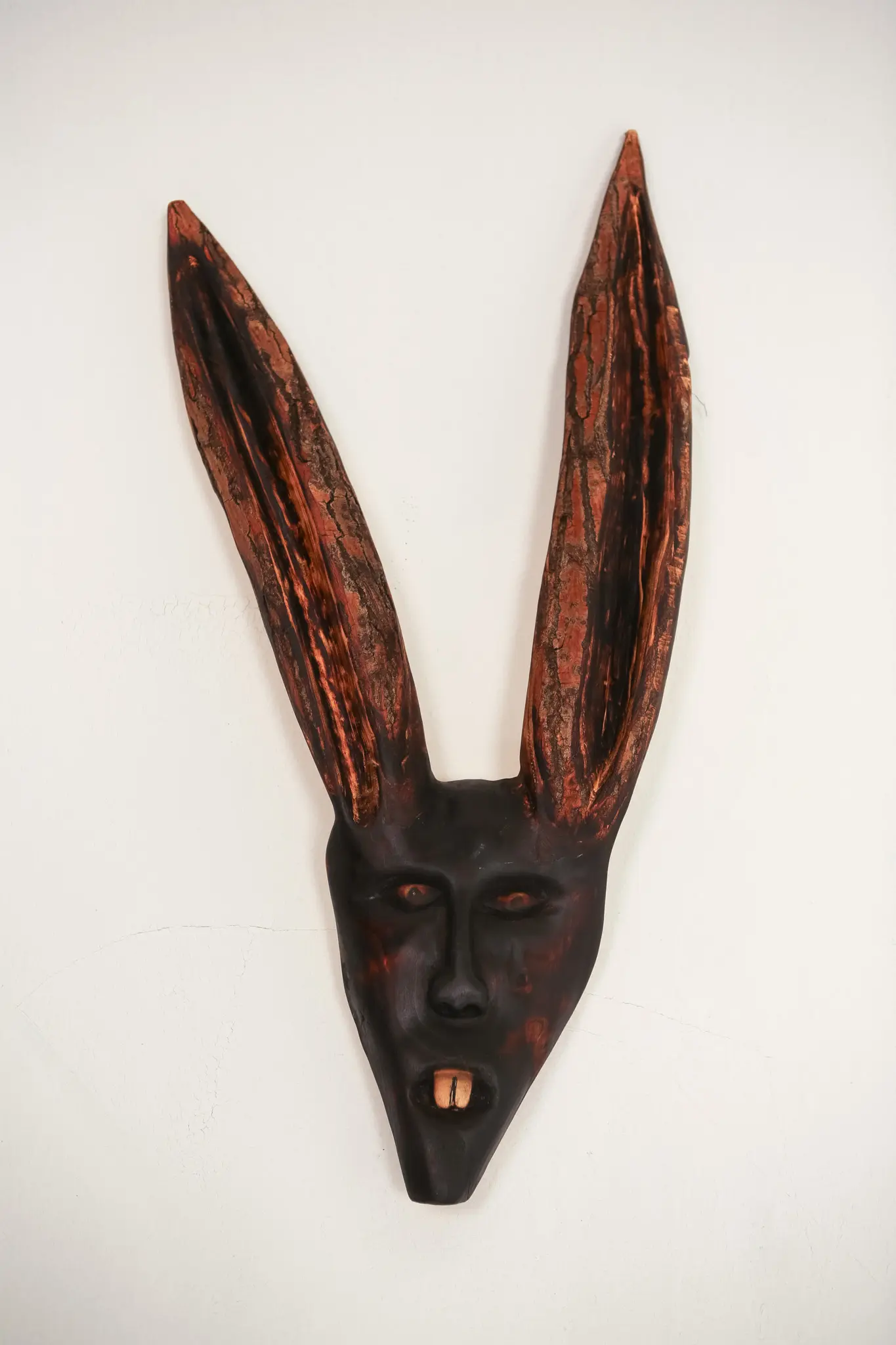 Même masque vu de face, oreilles verticales rapprochées, visage sombre aux reflets rouille et bouche entrouverte sur une dent pâle.
