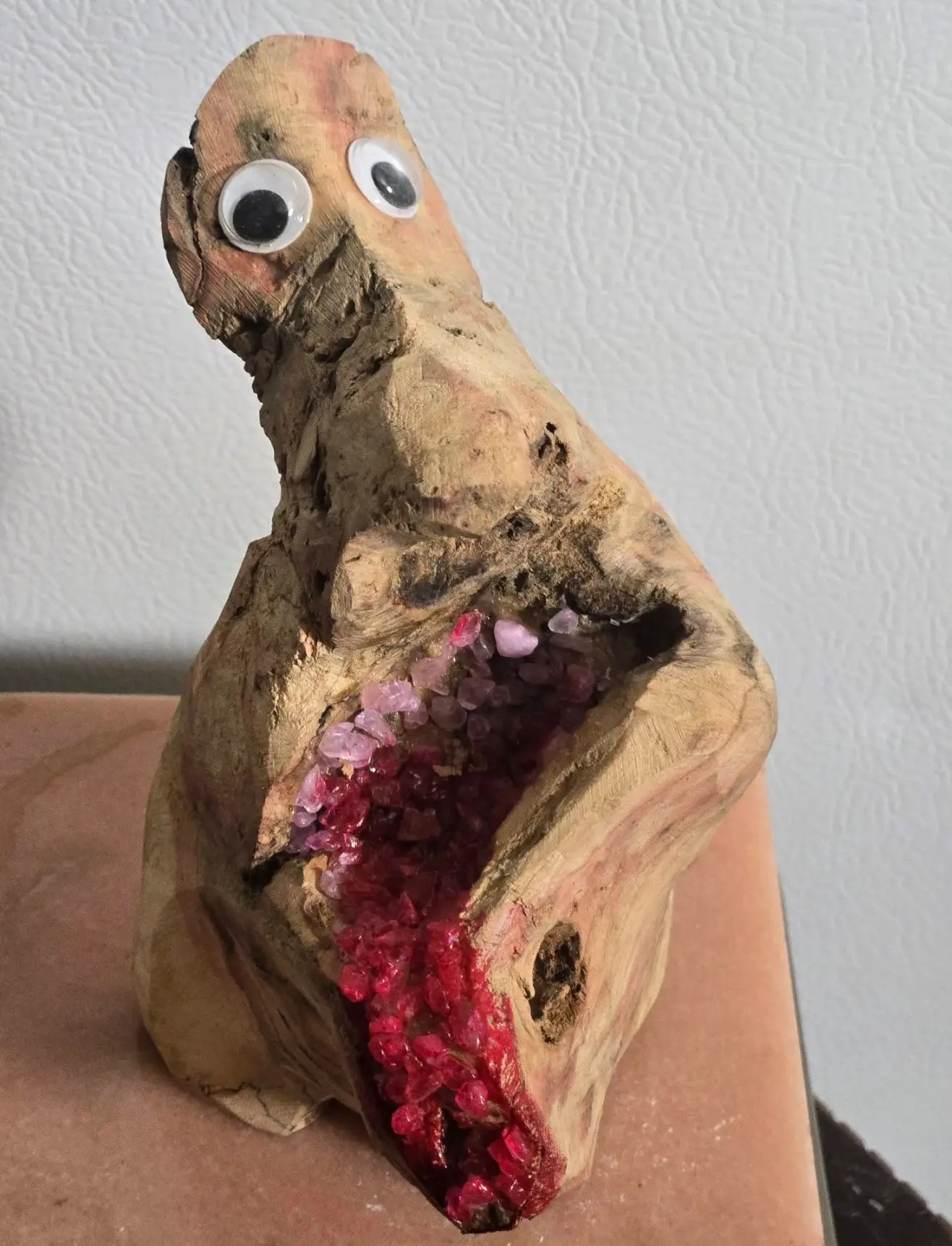 Sculpture en bois trouvé clair, deux yeux mobiles près du sommet, une large cavité sur le devant garnie de fragments translucides passant du rose pâle en haut au rouge soutenu en bas, un petit trou sur le flanc droit, posée sur un comptoir rosé devant un mur blanc, œuvre de Robert Bibeau
