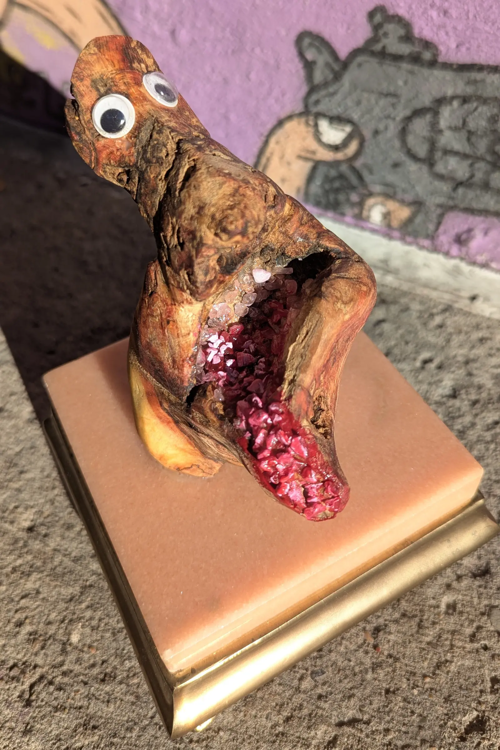 Bouche Bée vue de trois-quarts en plongée, les deux yeux mobiles vers l'objectif, la cavité garnie de fragments rose pâle puis rouge vif jusqu'au rebord, un lobe de bois blond au pied, socle de marbre rose à rebord doré, mur peint violet