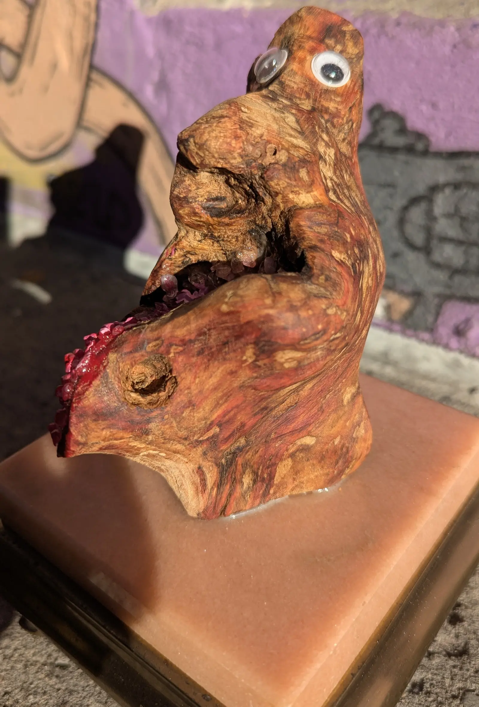 Bouche Bée de profil droit, la tête au sommet avec ses deux yeux mobiles, le bois veiné de rouge orangé et de plages pâles, le petit trou rond au flanc, le rebord rouge de la cavité au bord gauche, socle de marbre rose, mur violet