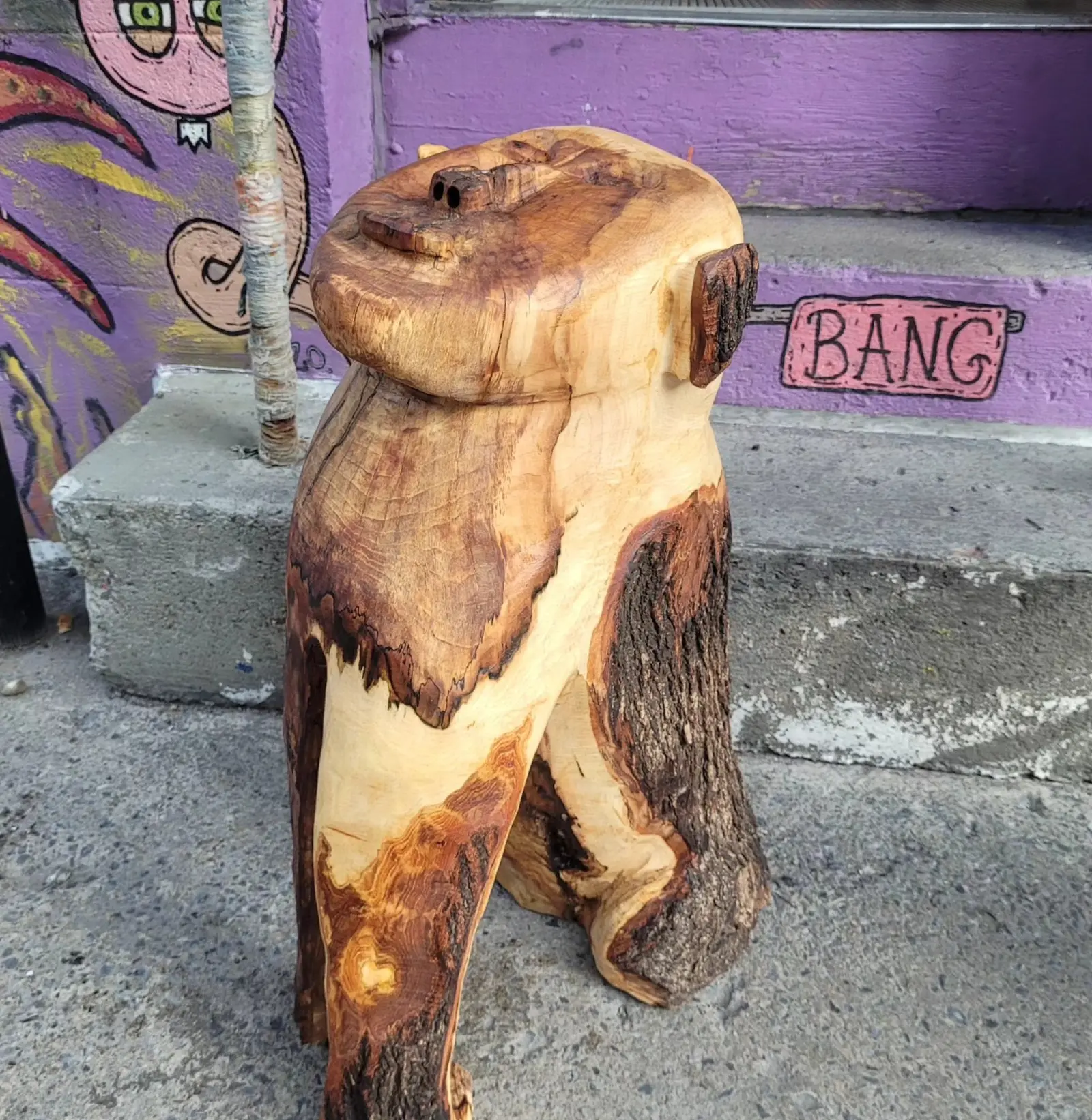 L'œuvre posée sur un trottoir de béton devant un mur violet couvert de graffitis, une marche et le tronc d'un jeune arbre sur le côté, tête renversée en arrière, écorce grise conservée le long de la jambe