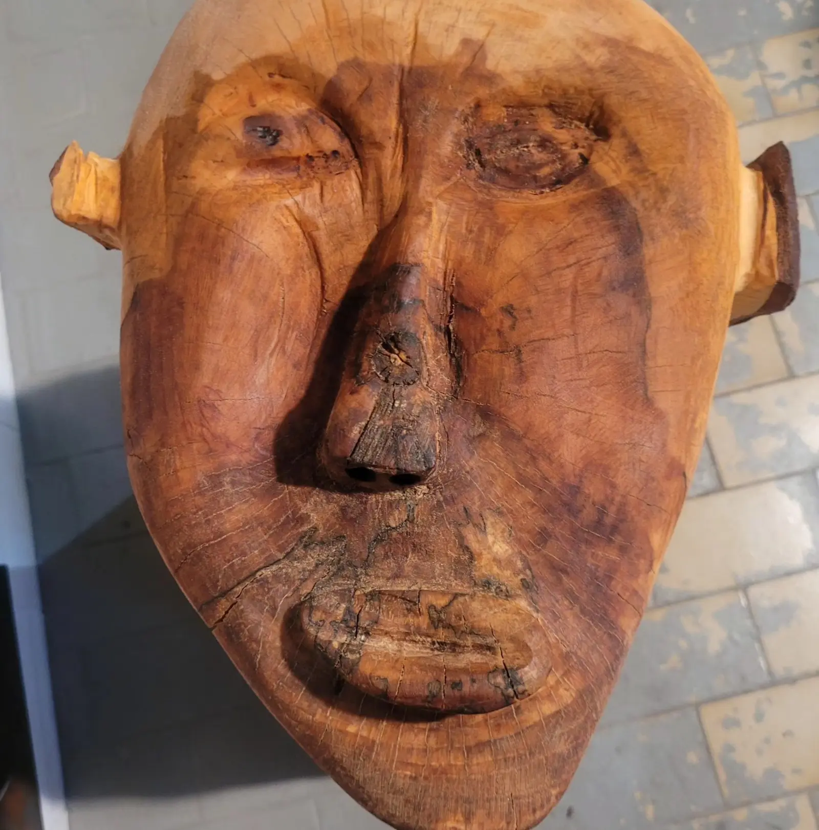 Gros plan du visage taillé dans le bois, yeux mi-clos en amande, nez saillant percé de deux narines, bouche en fente creusée dans une zone d'écorce sombre, une oreille carrée pâle à gauche et une oreille foncée à droite, sol carrelé flou derrière