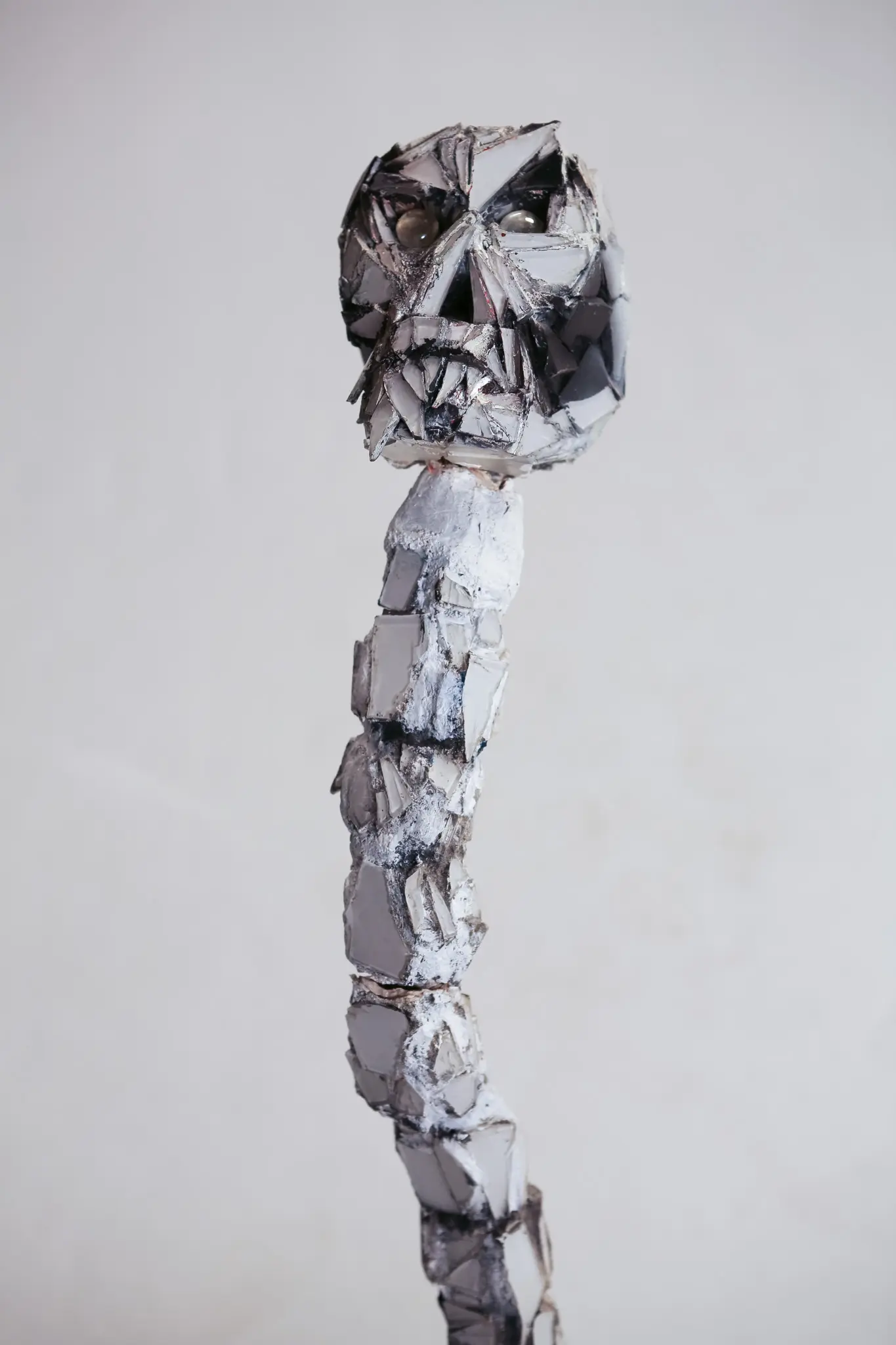 Figure verticale en eclats de miroir, crane de face aux orbites creuses garnies d yeux de verre, monte sur une colonne vertebrale segmentee, sur fond clair.