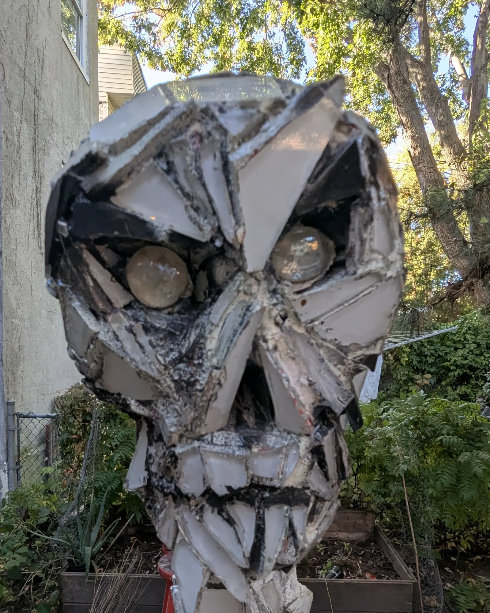Frontal close-up of the skull outdoors, mirror shards with a pale grey reflection set in dark grout, two domed amber glass eyes deep in the sockets, the top of the column below the chin, trees and a beige wall behind