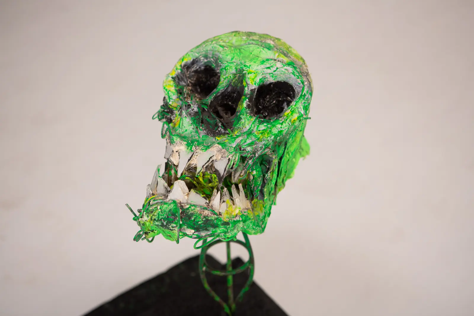 Crâne vert de face légèrement plongeante, deux orbites sombres et rangée de dents en verre brisé, posé sur socle noir.