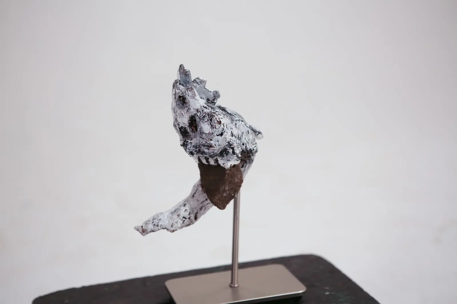 Vue d ensemble de la sculpture sur sa tige metallique et son socle, roche brute saillante a la base de la tete, fond clair.