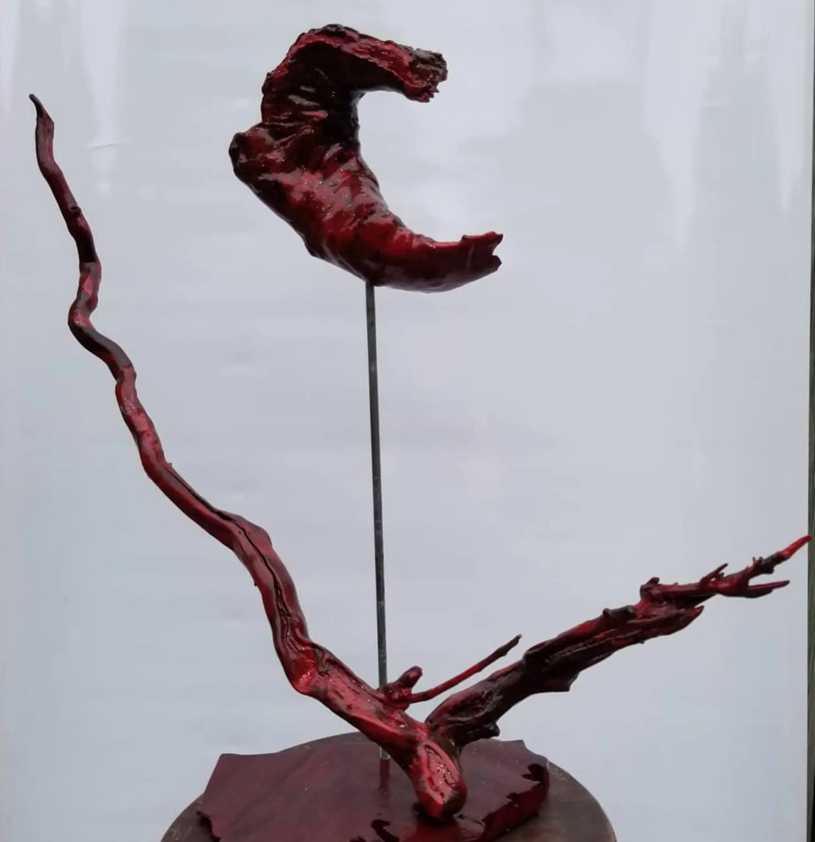 Sculpture rouge vif et brillante montée sur une base ronde en bois sombre : deux branches de bois trouvé s'ouvrent en V, et une forme creuse en croissant s'élève au bout d'une longue tige métallique, sur fond gris clair, œuvre de Robert Bibeau