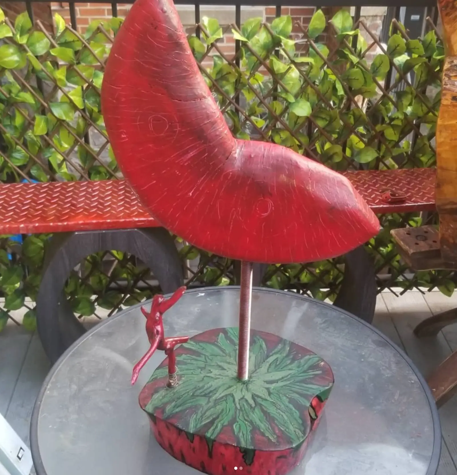 Croissant de bois peint en rouge, surface striée de rayures claires, monté sur une tige métallique filetée fichée dans un socle rond peint de feuilles vertes où se tient une petite figure rouge aux bras levés, œuvre de Robert Bibeau