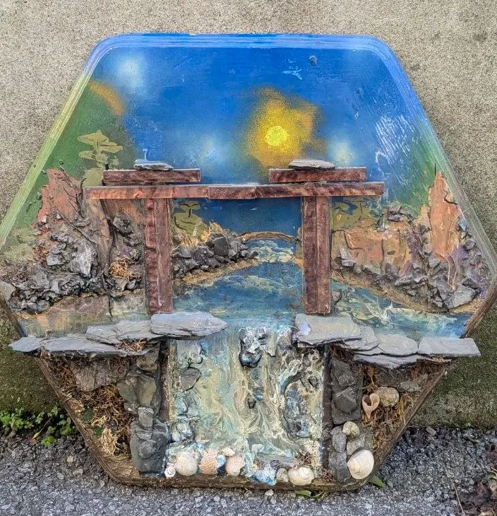 Diorama hexagonal en résine : un pont à portiques enjambe un ruisseau entre des berges de schiste et d'écorce, sous un ciel bleu au soleil tourbillonnant façon Van Gogh, cascade et coquillages incrustés à la base, œuvre de Robert Bibeau