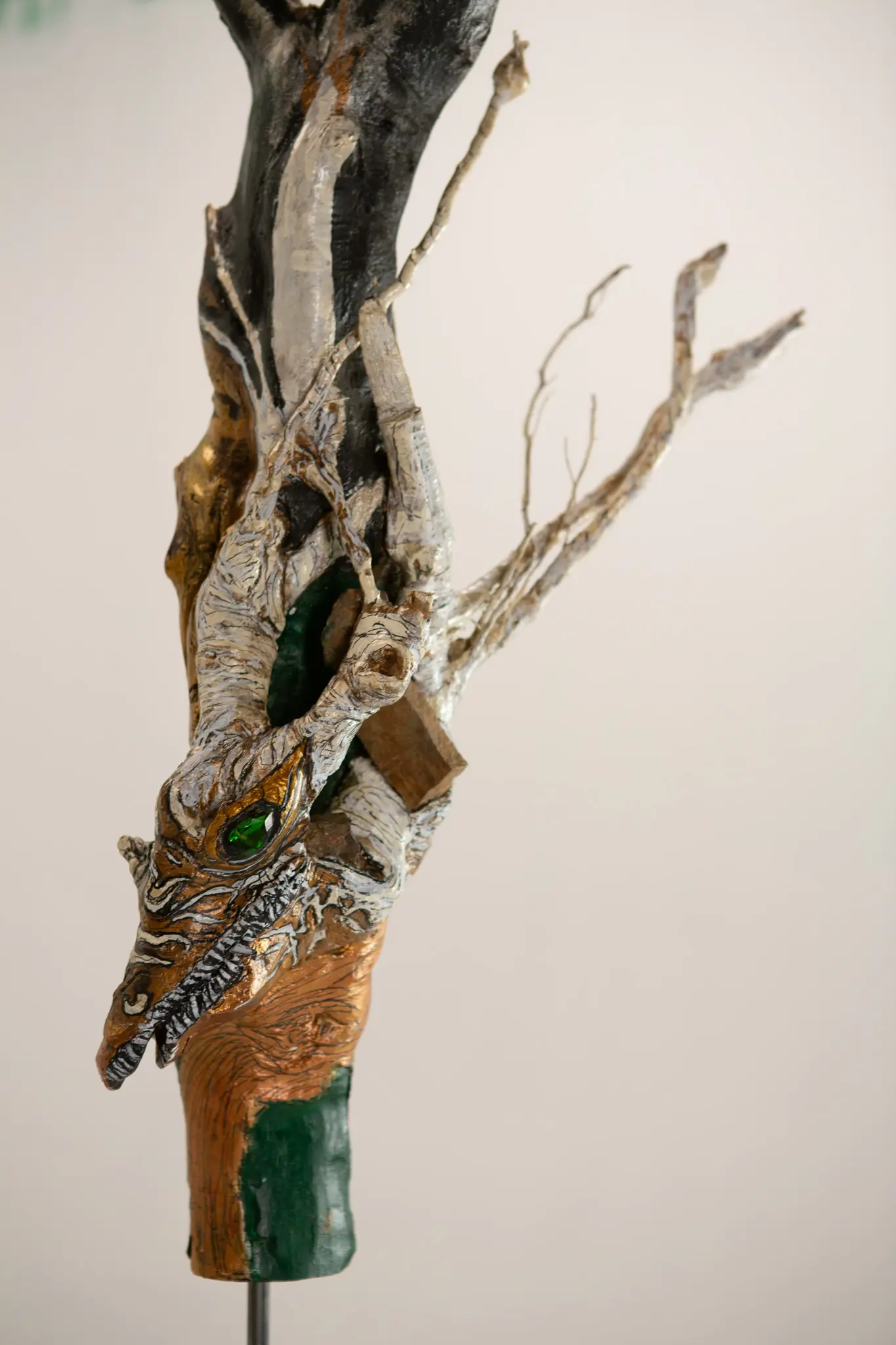 Sculpture verticale en bois trouve, tete de creature au museau allonge, oeil de verre vert, dorure a la feuille et branches pales dressees.