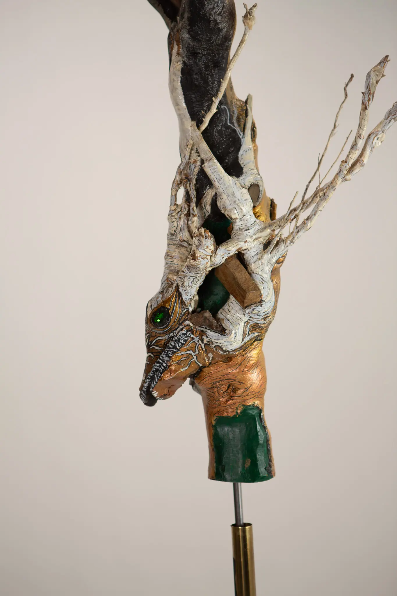 Tete de creature emergente au long museau, oeil de verre vert, branches pales et bloc de pigment vert a la base sur tige metallique.