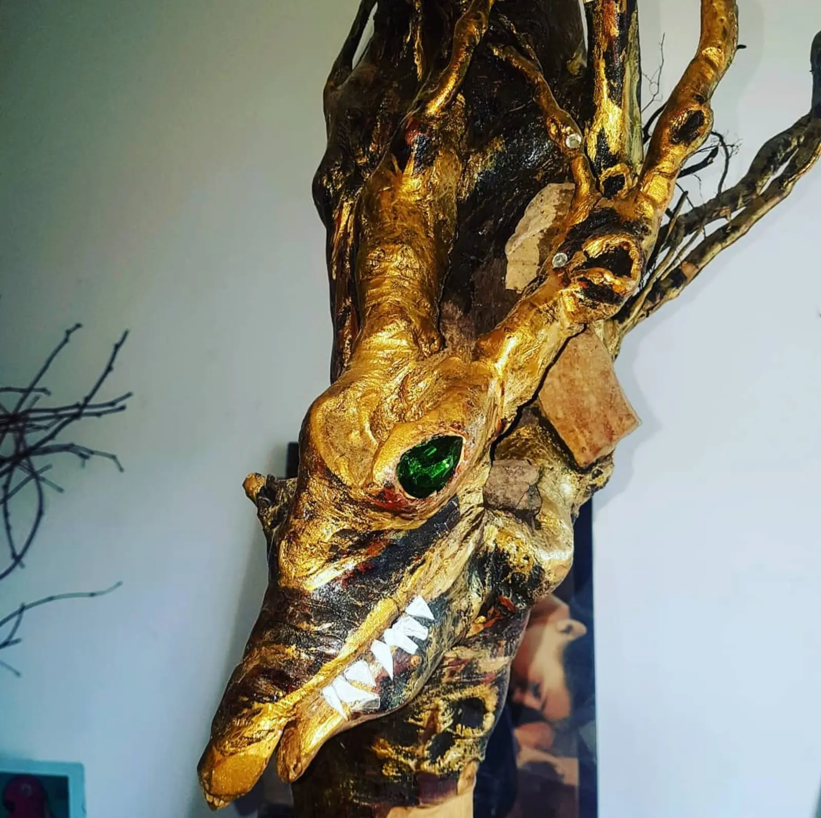 Close view of the gold-toned head, a green glass eye set into a hollowed socket and small white pointed teeth ranged along the jaw, against a pale wall where a spray of bare branches stands out