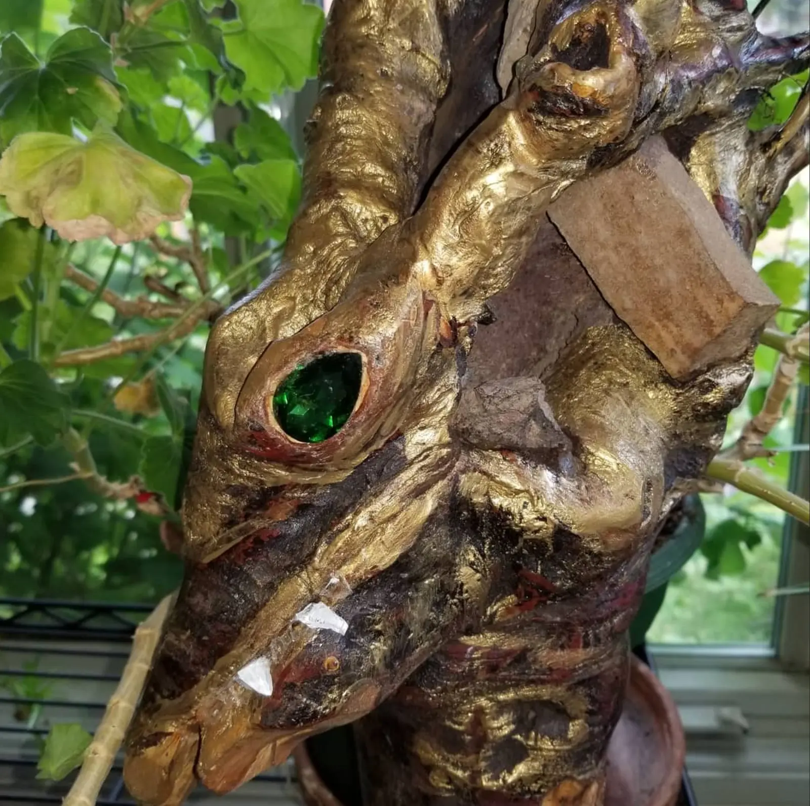 The gold-toned head seen up close in front of a window, a green glass eye in its socket, translucent white shards along the jaw and a pale wooden wedge driven into a split, potted foliage to the left