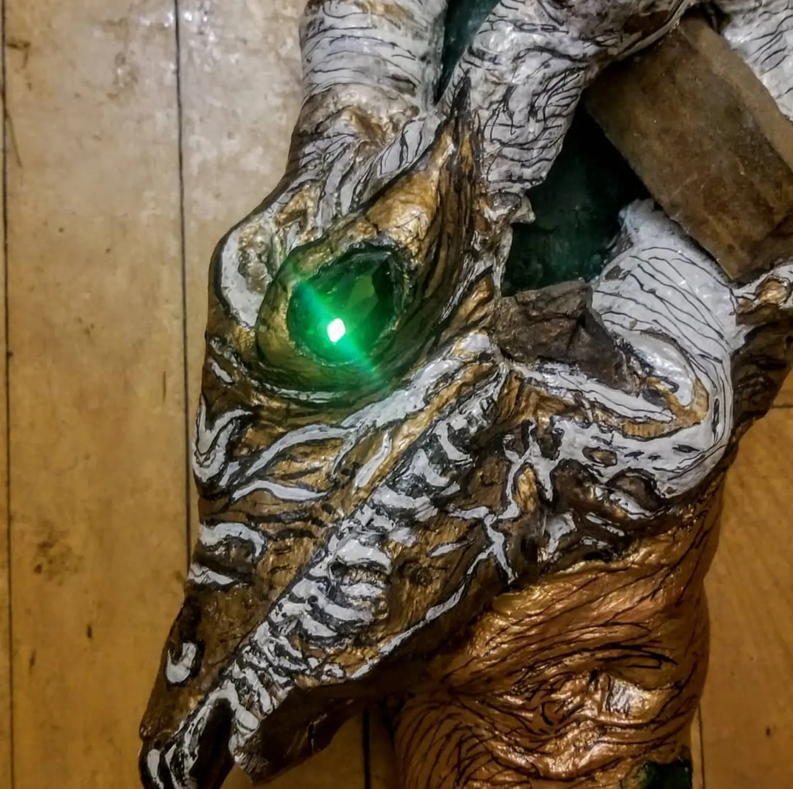 Detail of the head in white and gold with black linework, its green glass eye pierced by a point of light, a green painted hollow and a wooden wedge behind, against a wall of pale planks