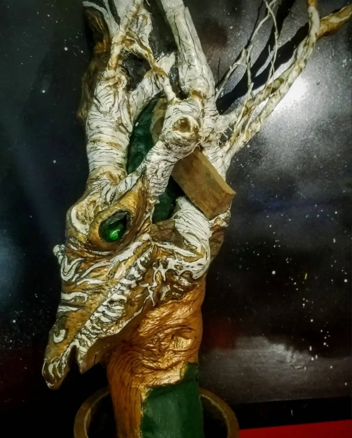 The work against a black backdrop flecked with white, its white and gold head carrying a long muzzle and a green glass eye, thin branches rising to the right, a red band running down the right edge of the frame