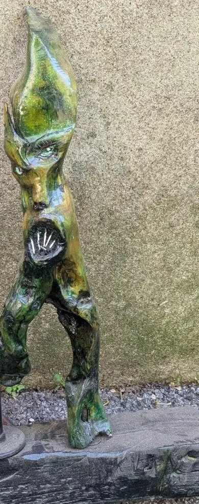 Sculpture verticale en bois sculpté à polychromie vert-de-gris et or, vue de face : un visage émergent aux yeux creusés et à la bouche en éventail clair, figure dressée sur ses jambes.