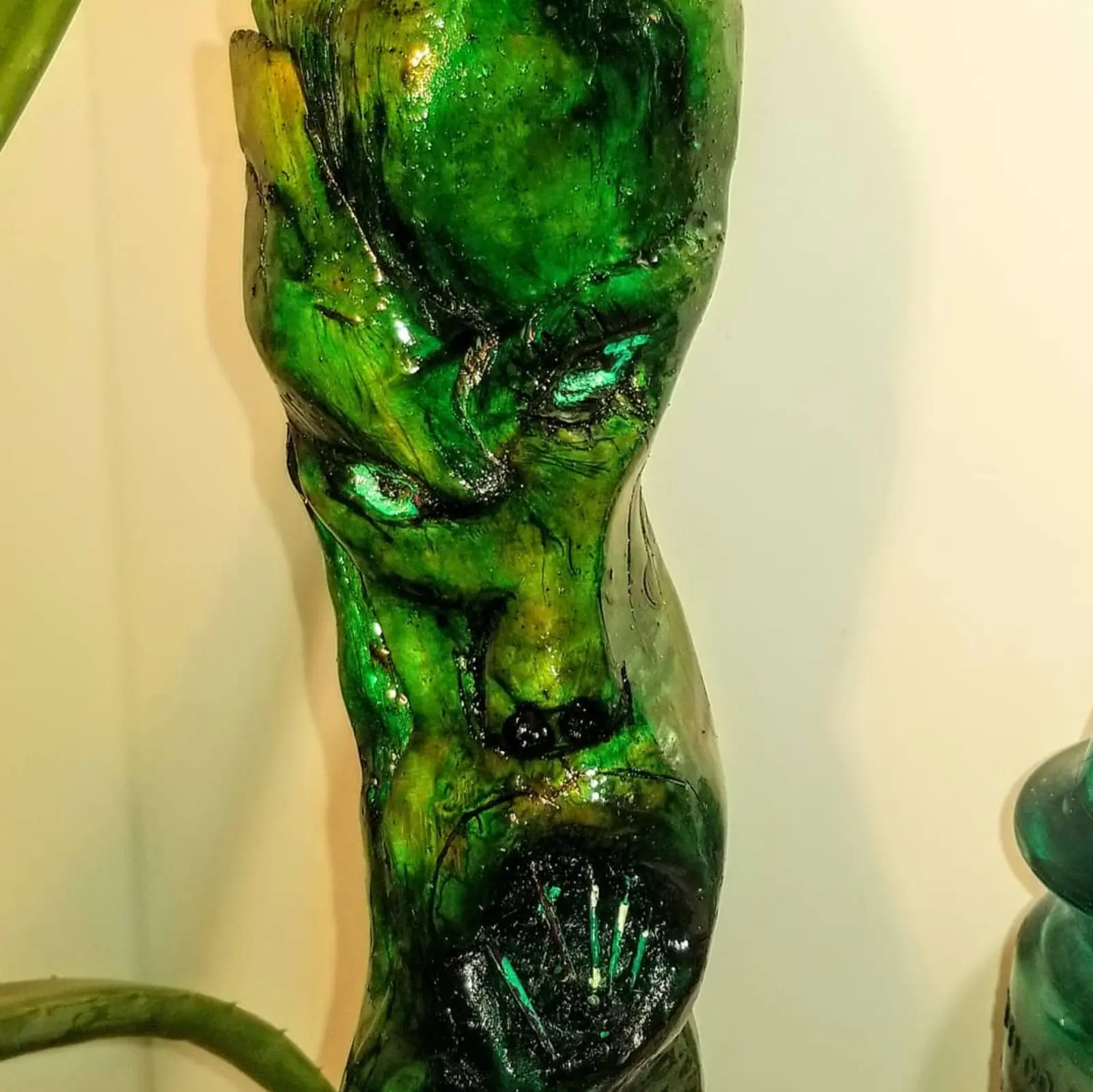 Gros plan sur le visage vert et luisant taillé dans le bois, l'œil mi-clos sous une arcade épaisse, deux petites boules noires en relief sous le nez et, plus bas, une cavité sombre hérissée de clous pâles, devant un mur clair