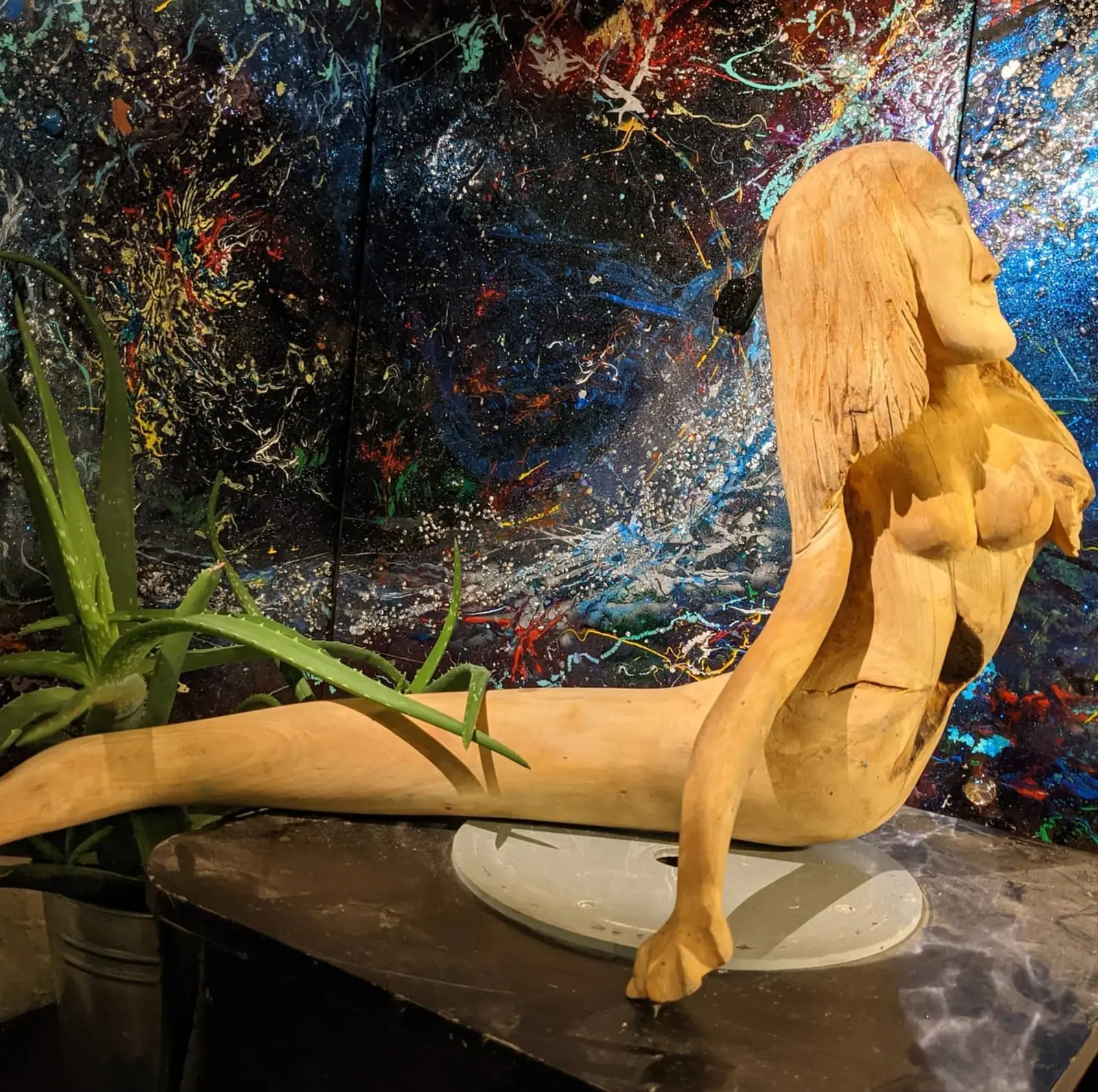 Sculpture en bois clair posée sur une table sombre, figure féminine nue vue de profil, buste redressé et membres allongés vers la gauche, longue chevelure striée dans le dos, un pied posé sur un disque ovale pâle, devant un panneau peint d'éclaboussures multicolores, œuvre de Robert Bibeau