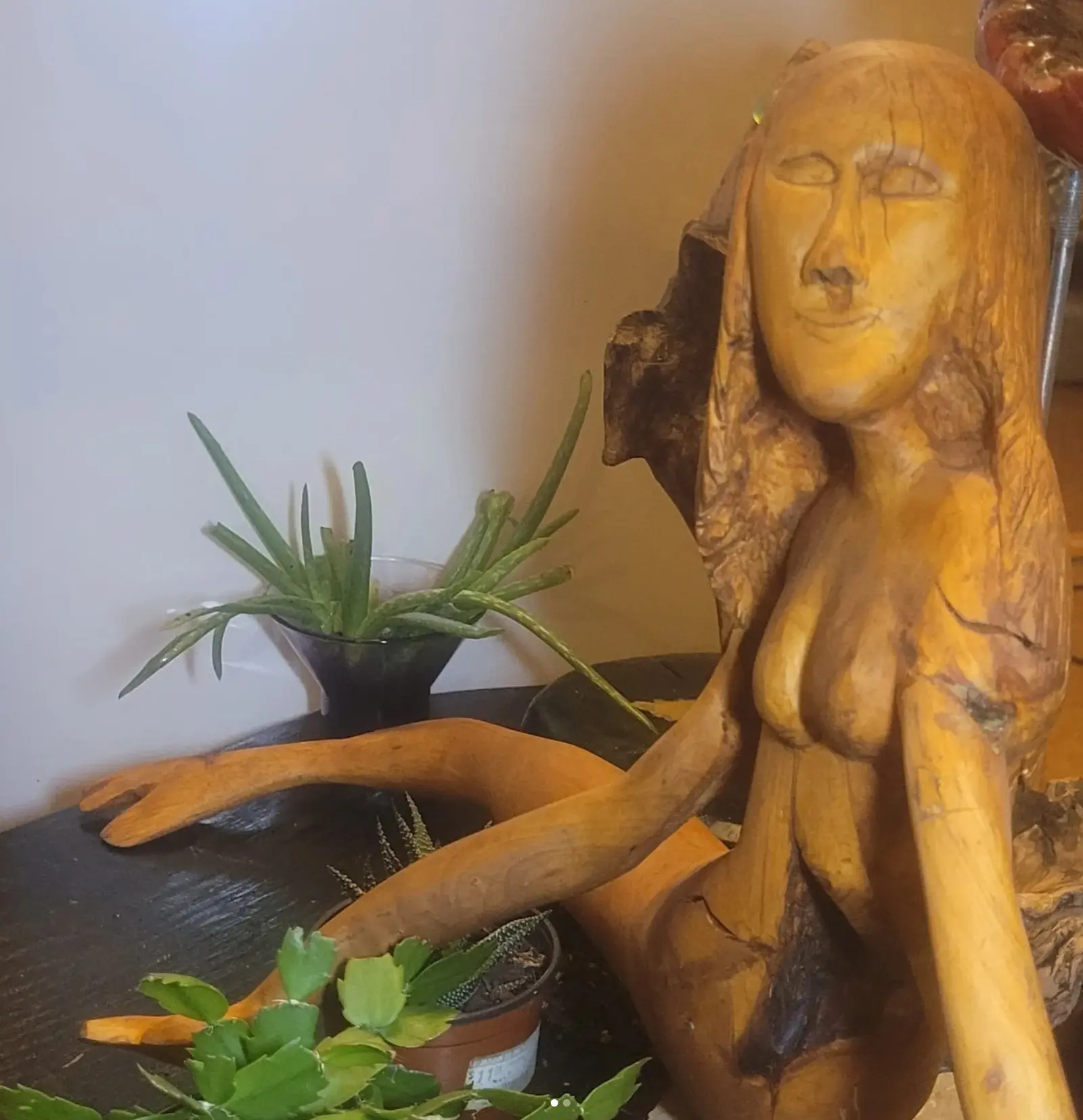 Vue rapprochée du visage et du buste en bois clair, yeux clos et bouche incisée, chevelure taillée en longues mèches, les bras allongés sur la table sombre et une main ouverte à plat, une masse de bois brut à écorce derrière l'épaule, plantes en pot et mur clair à l'arrière-plan.