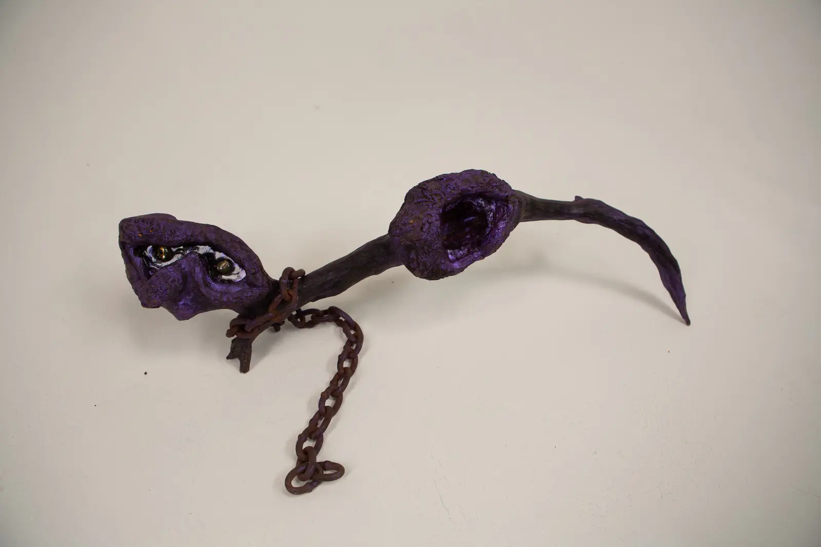 Sculpture horizontale en bois trouvé teinté de violet, vue d'ensemble, tête à gauche et cavité béante à droite, chaîne rouillée pendante.