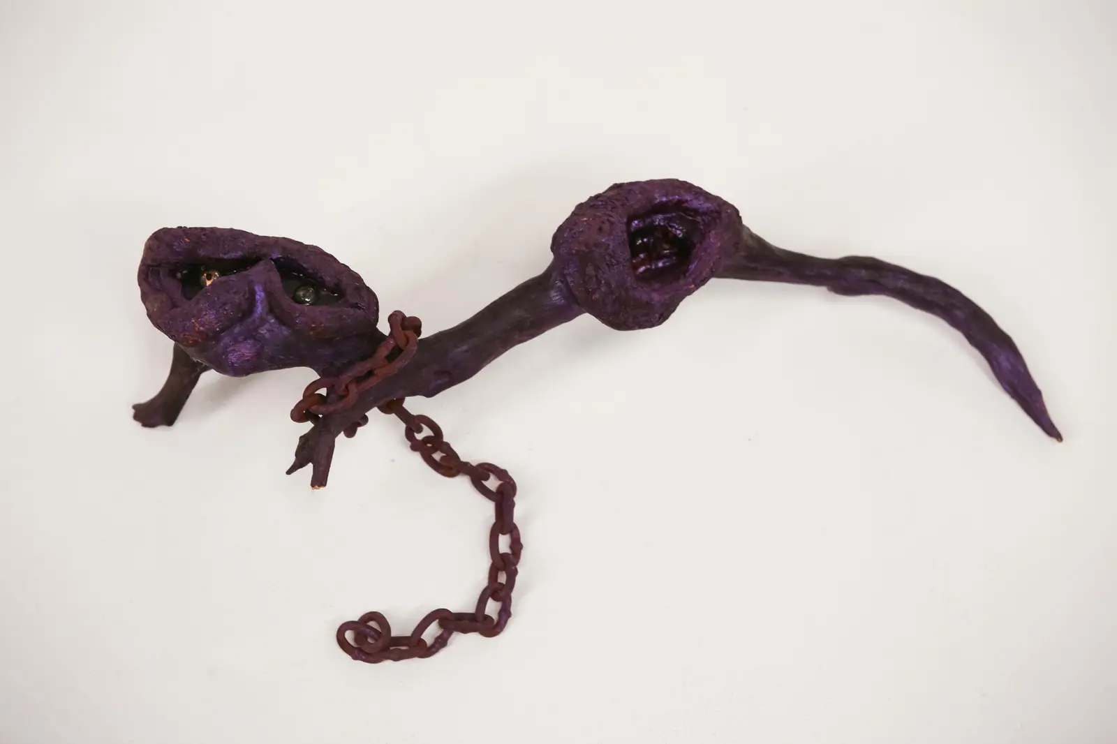 Vue de trois-quarts de la figure animale violette posée sur ses pattes fourchues, chaîne rouillée enroulée autour de la branche.