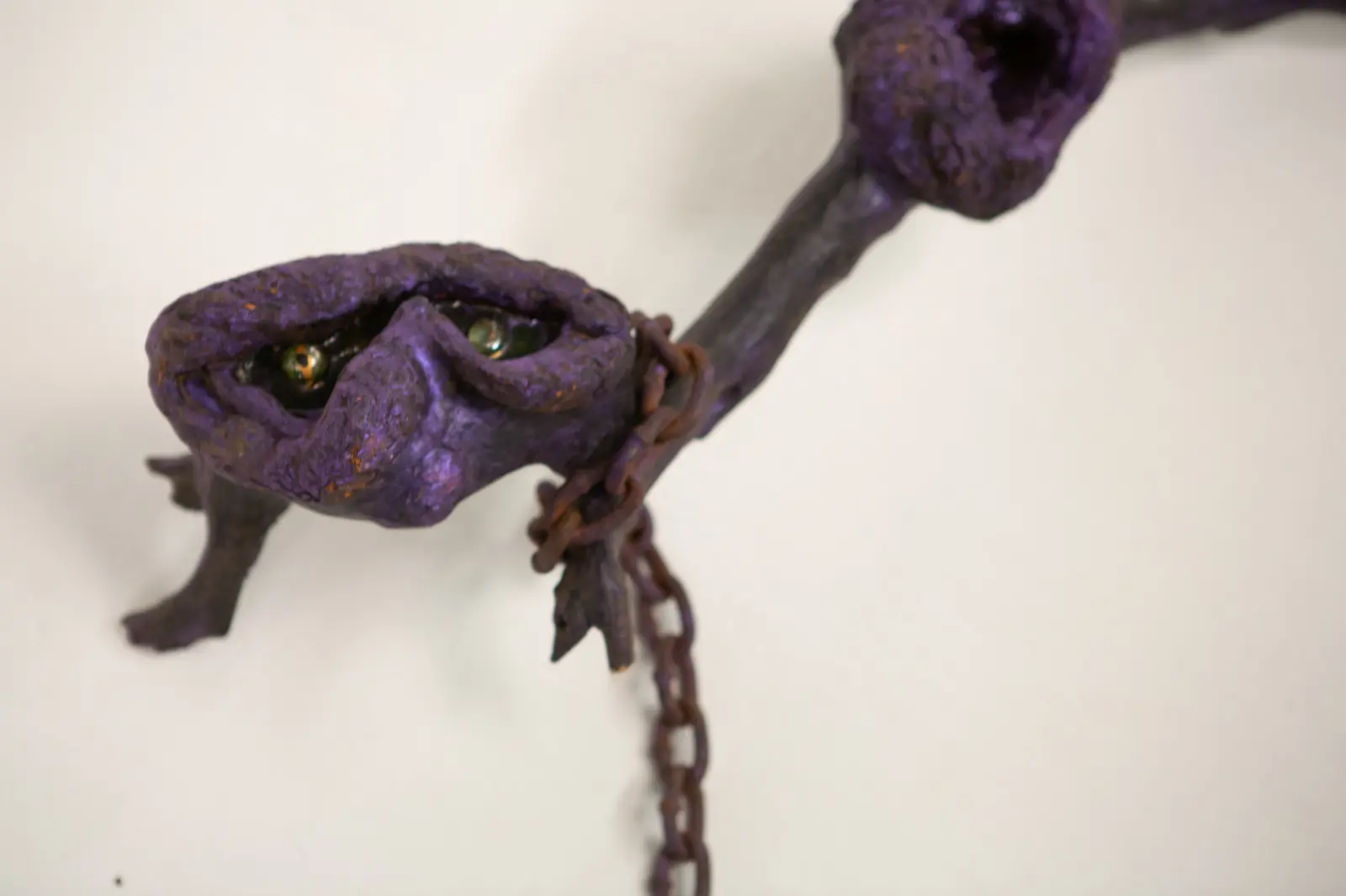 Gros plan sur la tête de la figure, deux yeux de verre logés dans une fente entre des bourrelets violets, chaîne au cou.