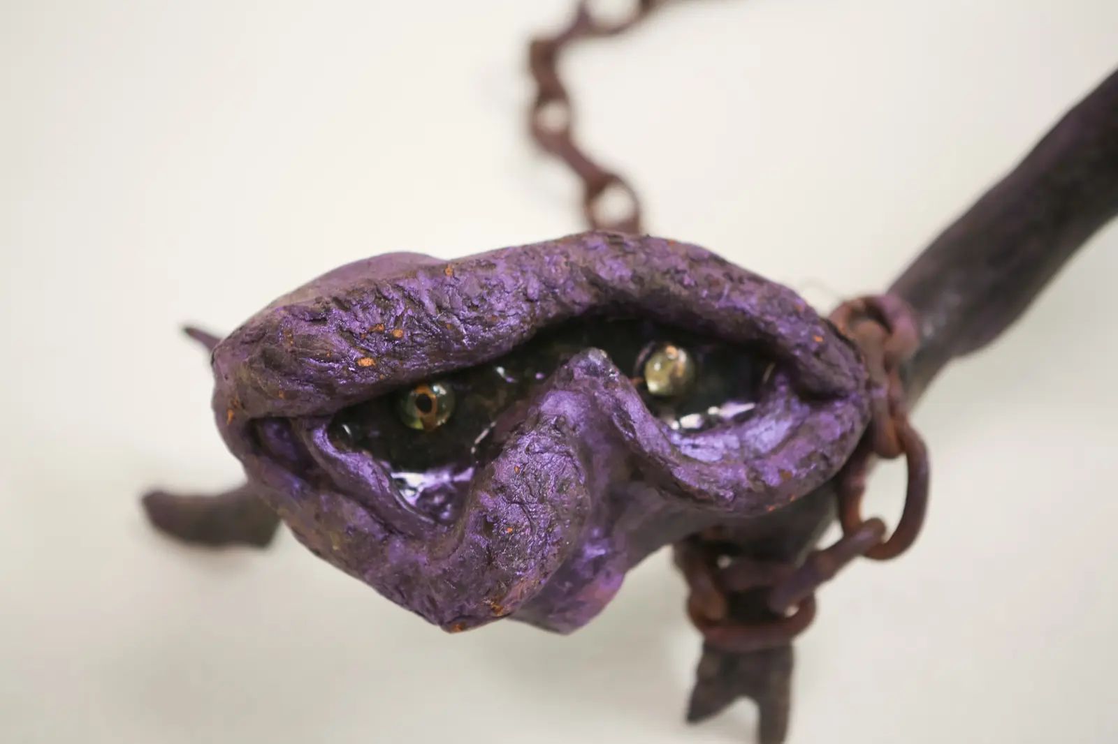 Plan serré frontal de la tête, deux yeux de verre ambrés derrière une fente bordée de bourrelets violets, maille de chaîne à droite.