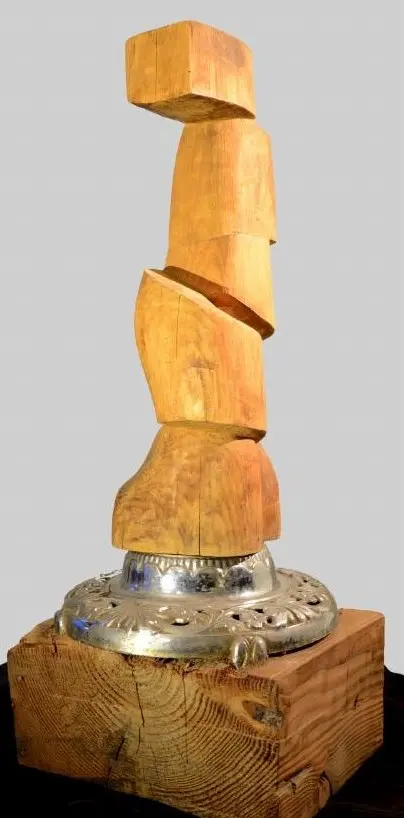 « Le boss » : colonne de blocs de bois empilés et vrillés sur une base argentée ornée, posée sur un bloc de bois brut, œuvre de Robert Bibeau