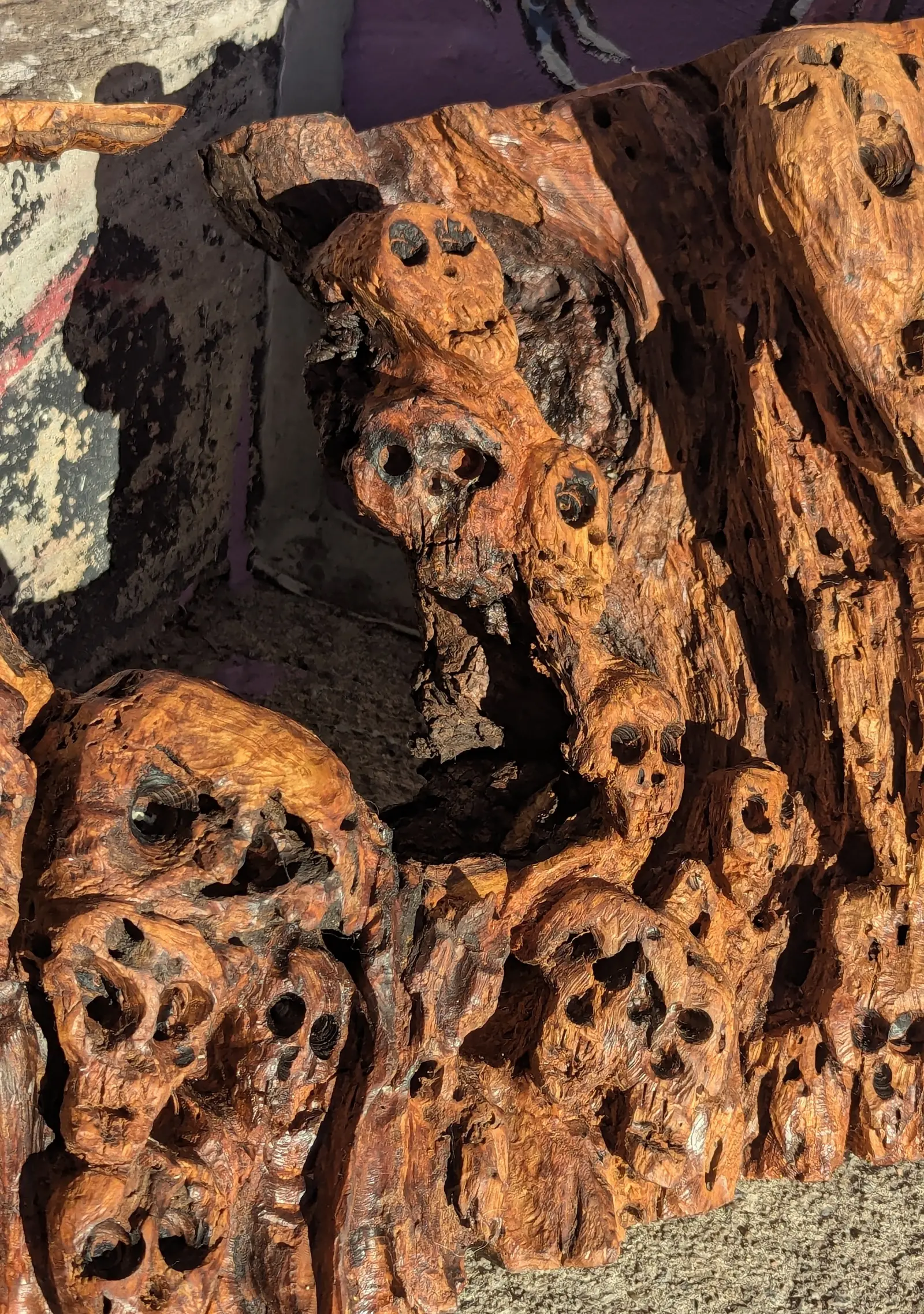 Mort mais vivant 2, gros plan sur les crânes du bas : petits crânes en grappe, orbites et nez en creux, un plus grand à gauche, deux autres en colonne au-dessus, bois orangé piqué de petits trous sombres, le creux profond du panneau en haut à gauche