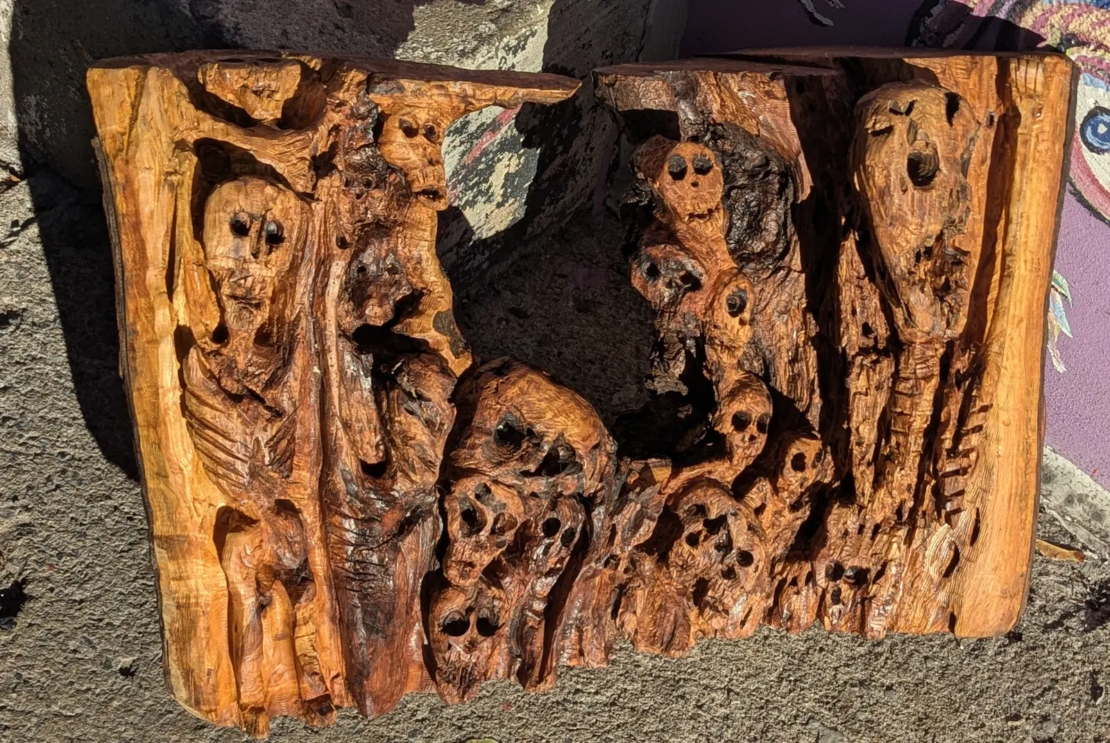 Mort mais vivant 2 : panneau de bois orangé à bords bruts, sculpté en bas-relief, une grande figure squelettique debout à gauche, des crânes empilés au centre, un long crâne creusé à droite, posé sur le béton devant un mur peint, œuvre de Robert Bibeau