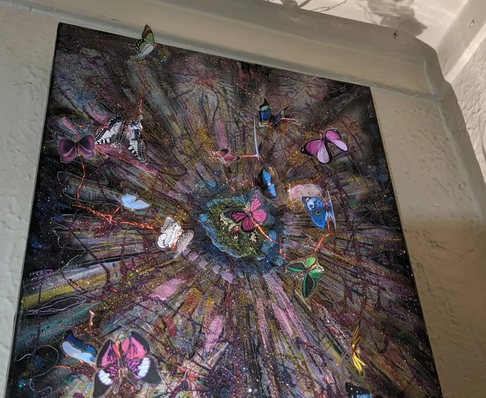 Tableau encadré accroché dans l'angle d'un mur blanc et du plafond, une dizaine de papillons colorés en relief autour d'un centre vert pailleté, sur un fond sombre à rayons violets et dorés, œuvre de Robert Bibeau