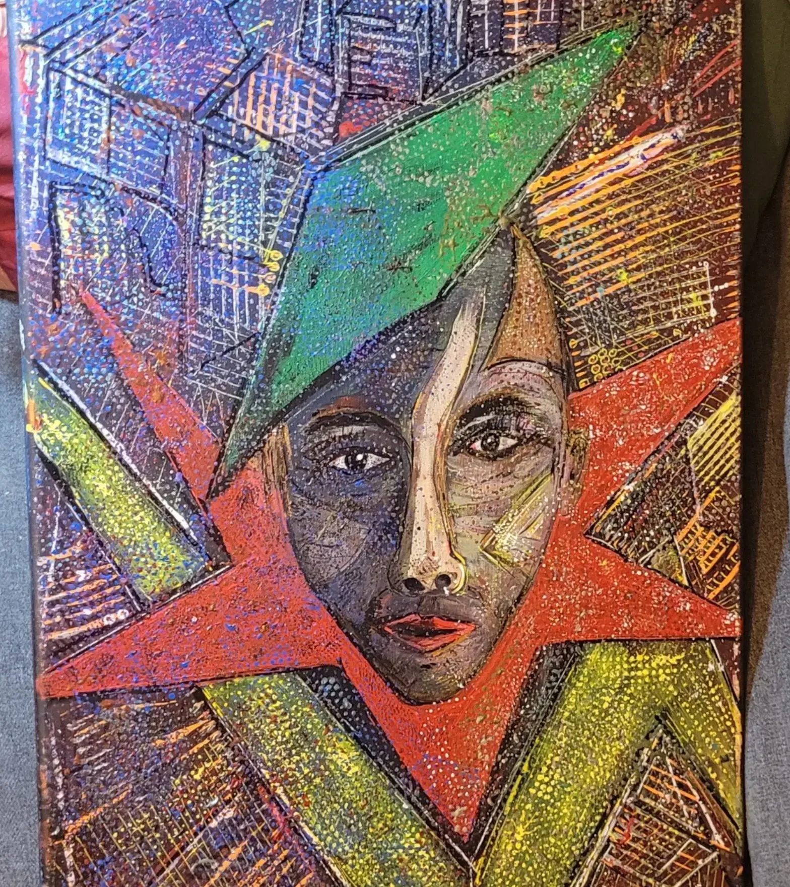 Toile appuyée de biais sur un sol gris, cadrage qui coupe le bord supérieur : un visage aux yeux grands ouverts, moitié grise et moitié ocre, occupe le centre d'une étoile rouge, sous un triangle vert et des grilles bleues