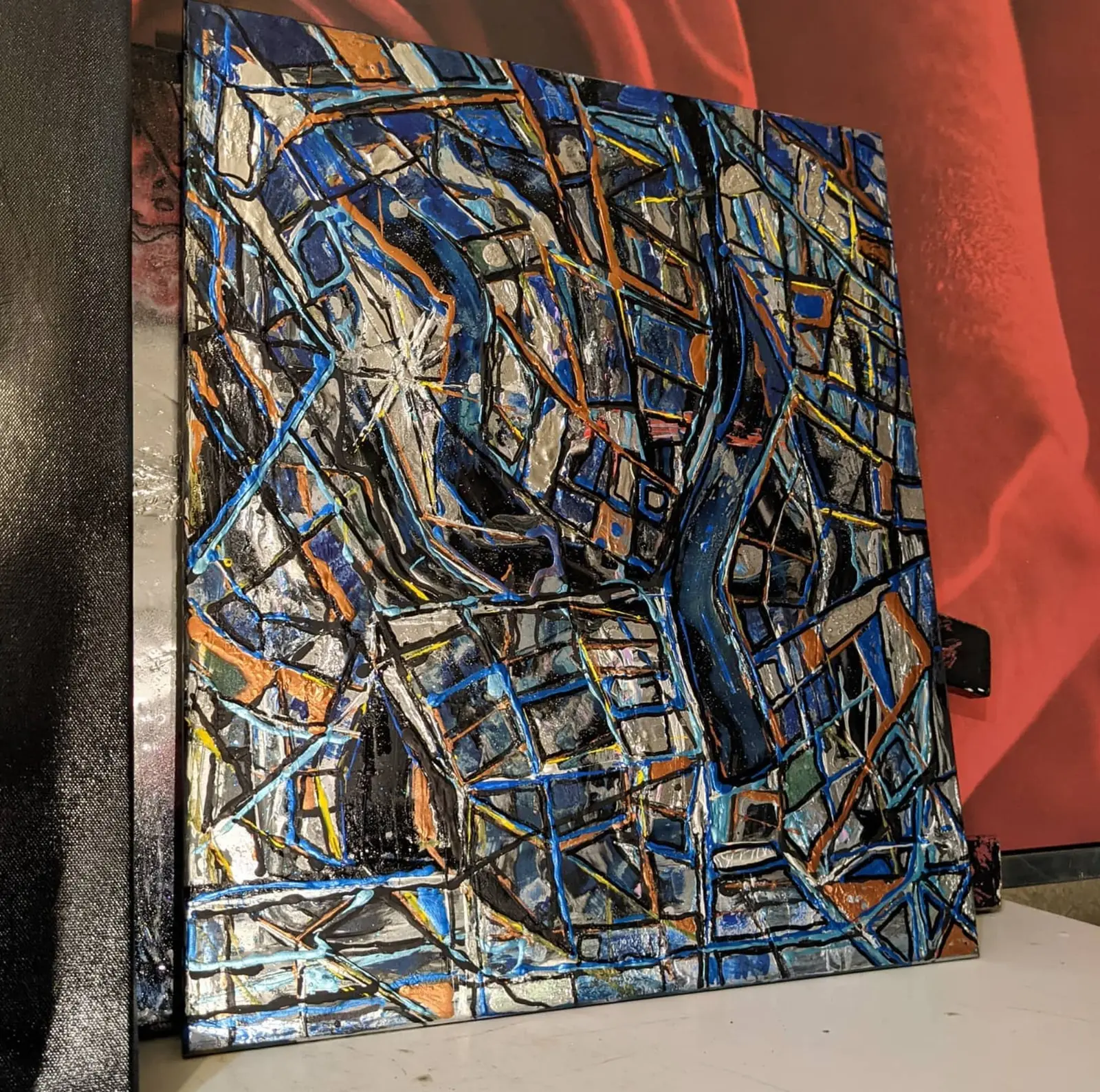 Tableau vu de trois quarts, posé sur un plan blanc contre une tenture rouge, surface en relief de lignes noires, bleues et orange sur gris argenté, une étoile blanche rayonnante vers le centre gauche