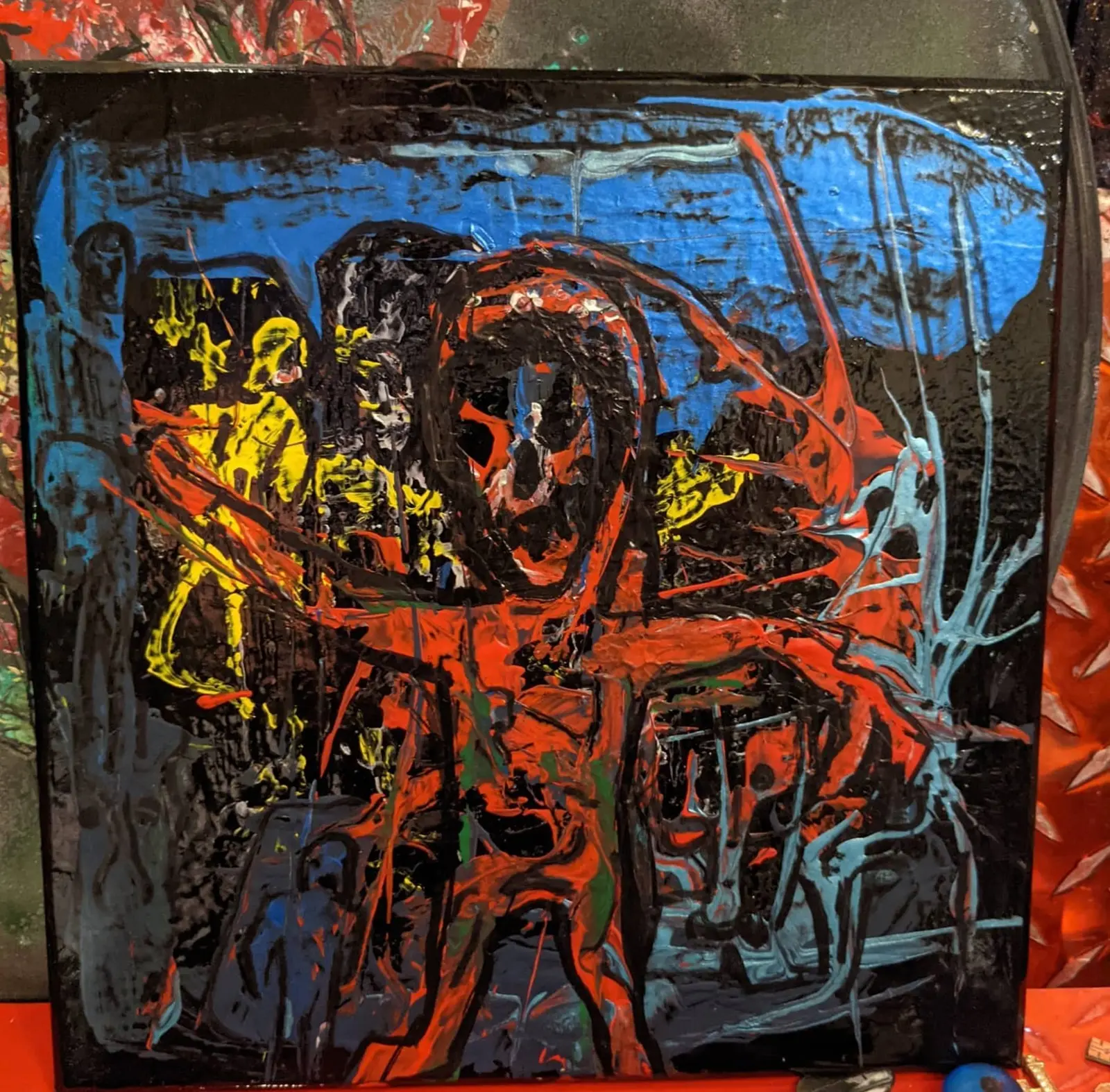 Toile carrée appuyée contre un mur d'atelier peint, matière épaisse et luisante : silhouette rouge aux bras ouverts, tête réduite à une masse noire cernée de rouge, figures jaunes à gauche, bande bleue en haut