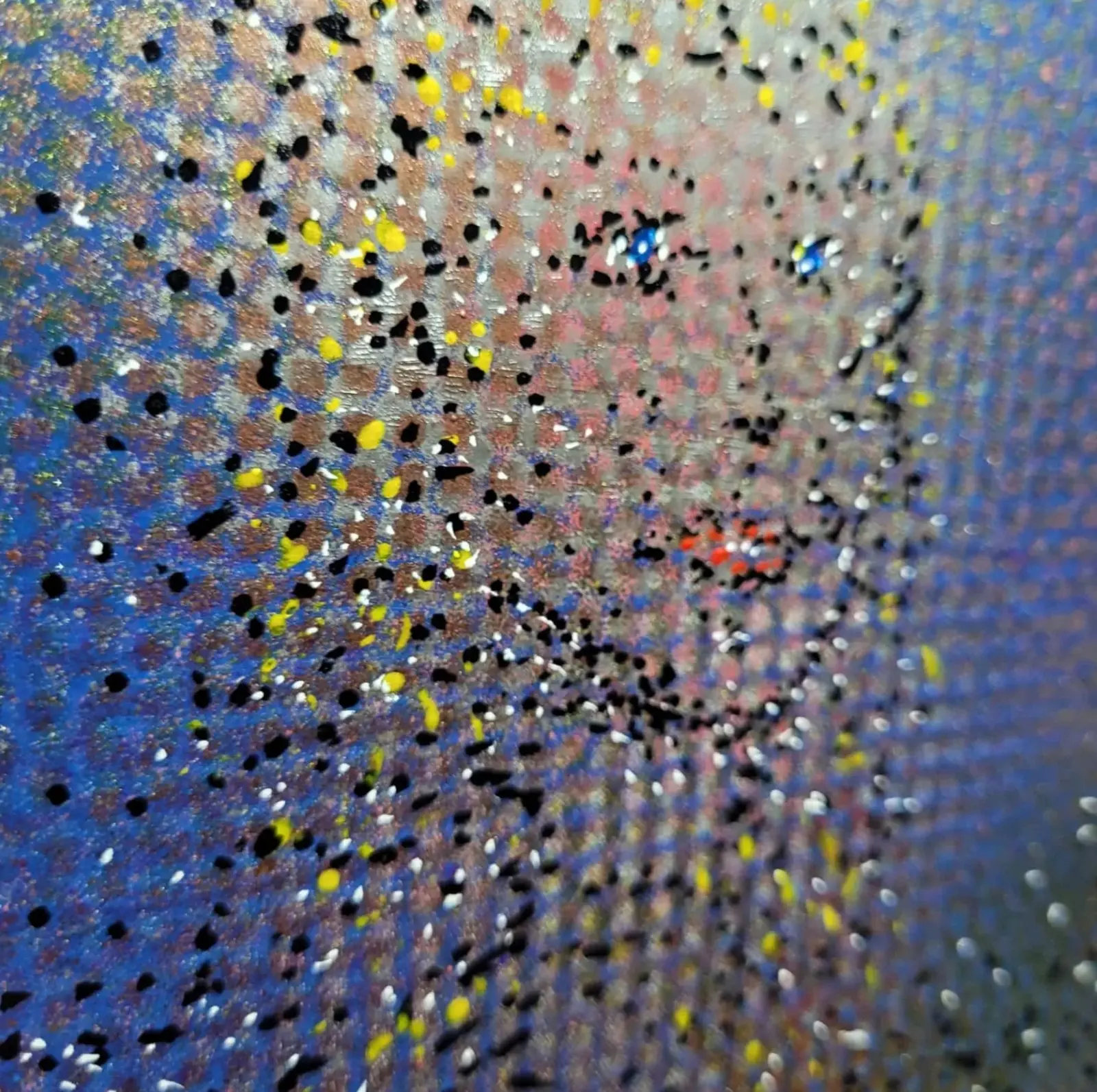 Gros plan en lumière rasante sur la trame de points d'une peinture, pastilles noires et jaunes épaisses semées de blanc sur un fond bleu quadrillé, deux points bleus et des traits rouges y dessinent des yeux et une bouche, bord droit flou