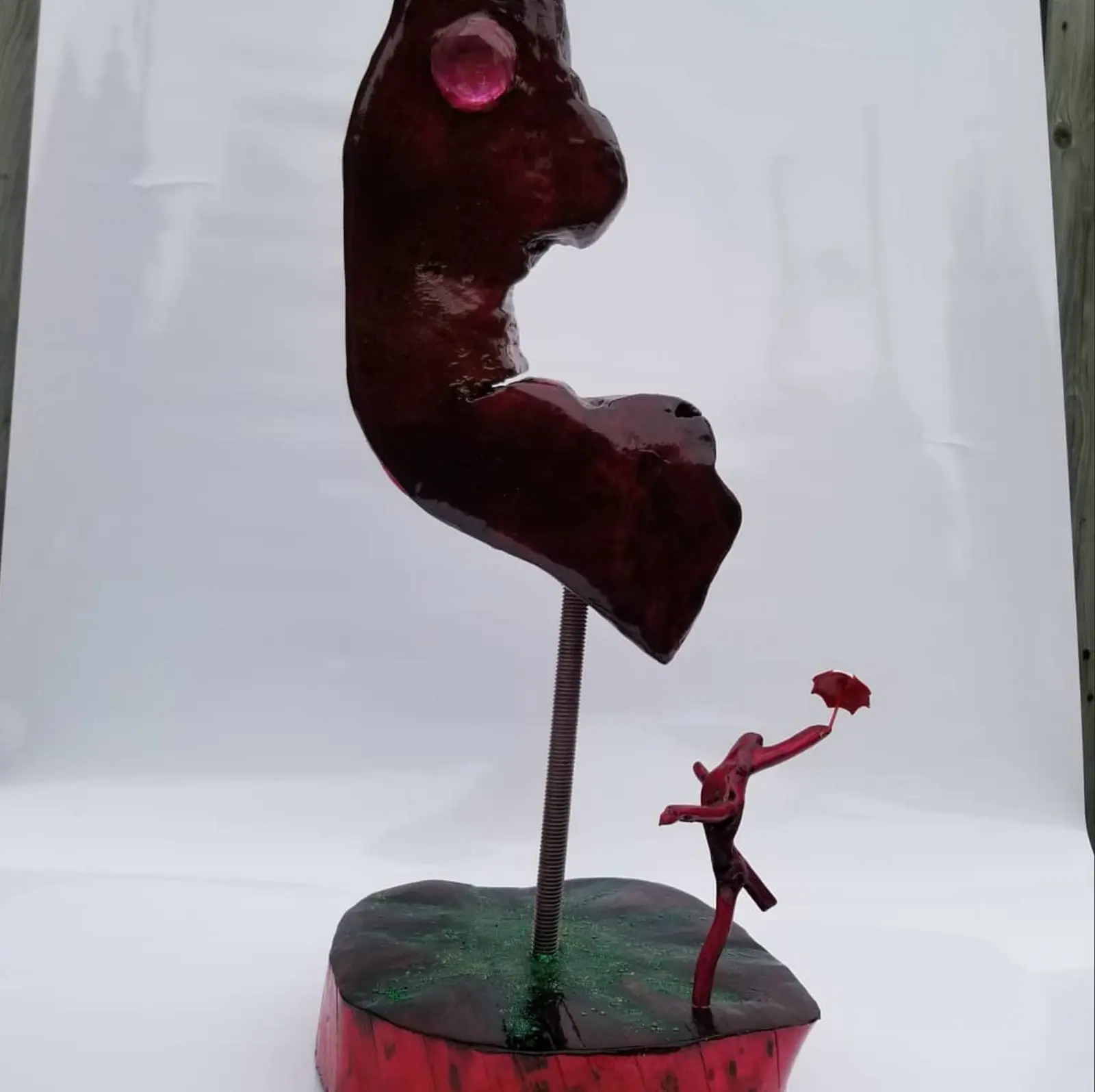 Haute forme de bois trouvé rouge sombre et vernie, sommet hors cadre, bille rose incrustée près de la cime, montée sur une tige filetée dans un socle en rondin rouge et vert où une petite figure de branchages tient un parapluie, œuvre de Robert Bibeau