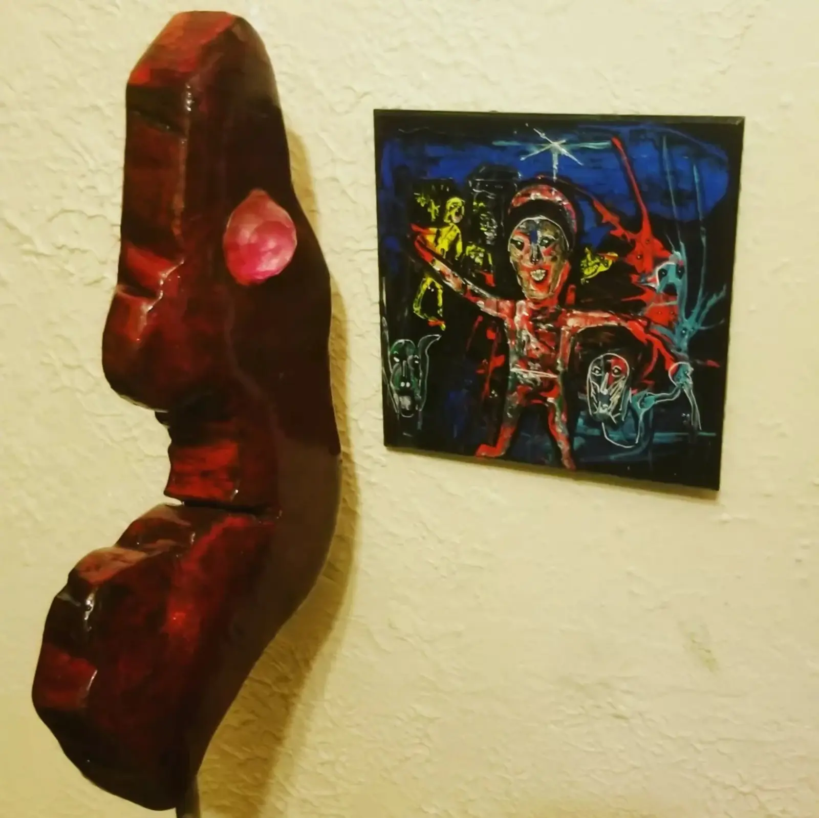 Sculpture rouge sombre vue de près en intérieur, cadrée sans son socle, la bille rose sur le flanc, dressée devant un mur crème texturé où elle porte son ombre, à côté d'un petit tableau carré bleu nuit à figure rouge et blanche