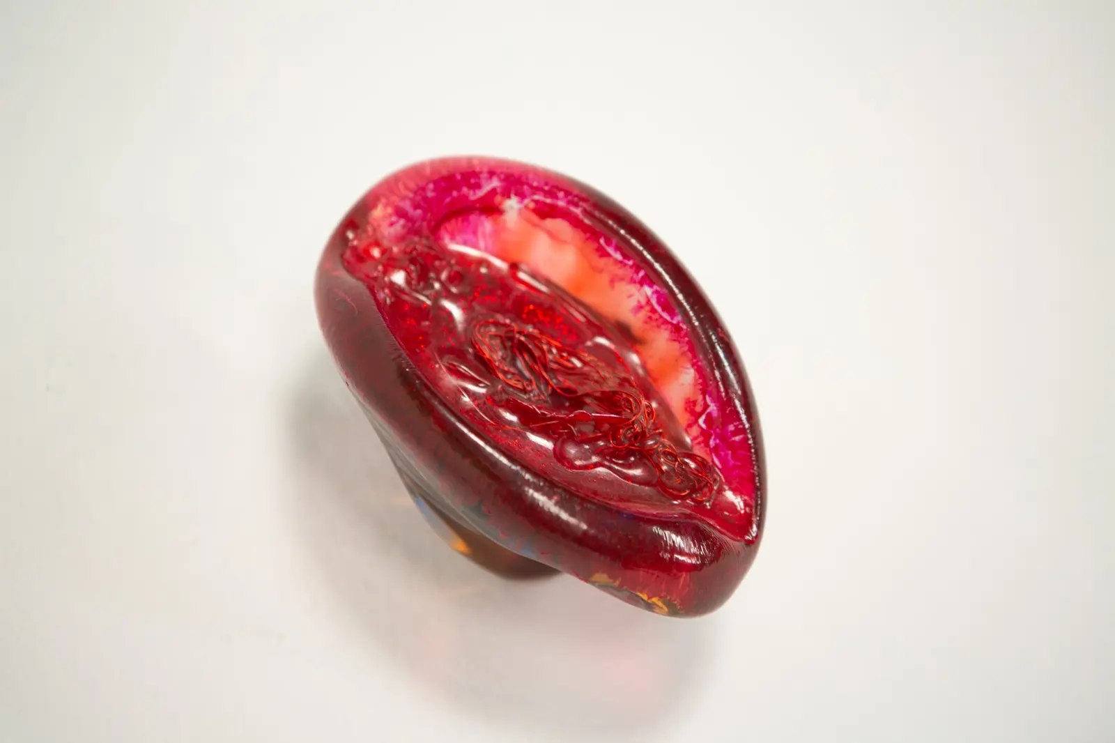 Forme ovoide en resine rouge translucide vue de trois-quarts, cavite centrale glitteree avec fil rouge enchevetre, sur fond clair.